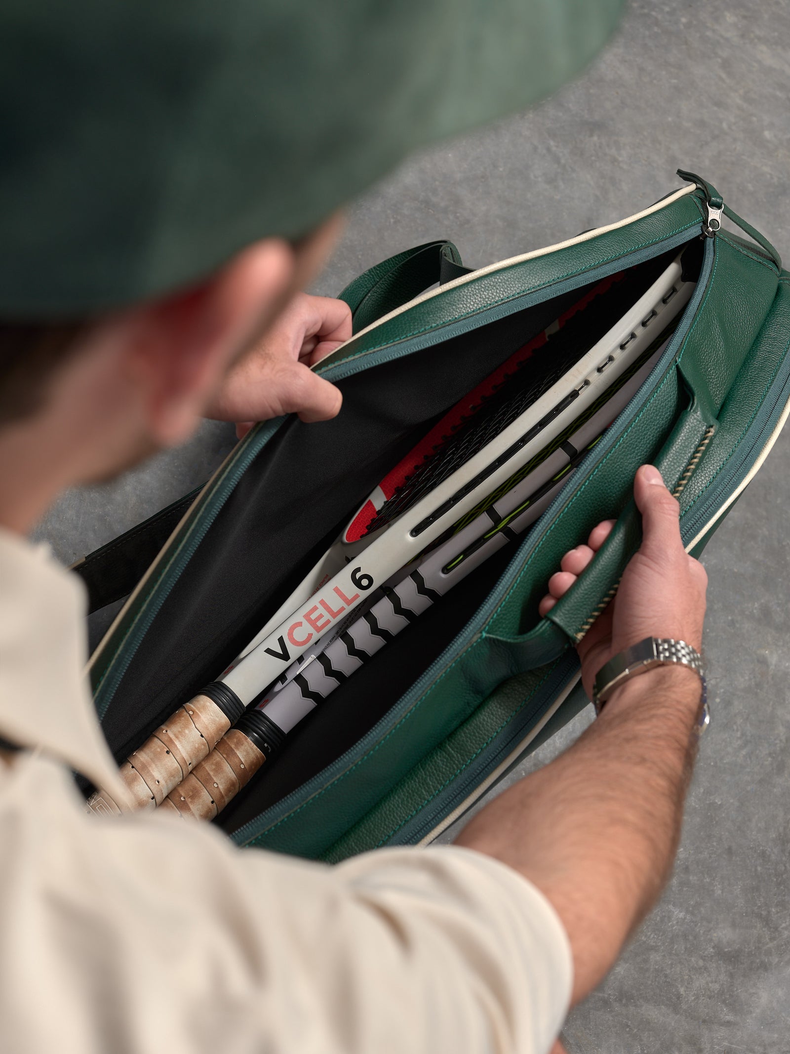 An overhead view into the open main compartment of the green leather tennis bag, showing two modern tennis rackets, the VCell 6 logo, and two leather-wrapped grips.