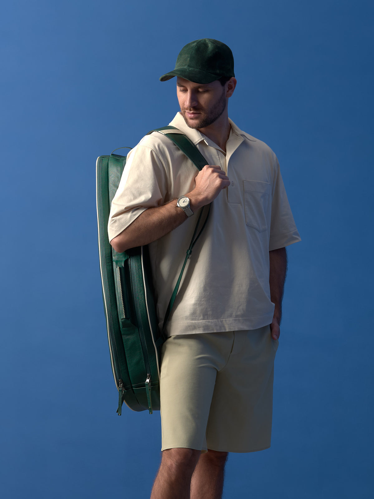 A man in a beige polo and green suede cap carries the handcrafted dark green leather tennis bag over his shoulder by its adjustable leather strap, showing the polished metal buckle.