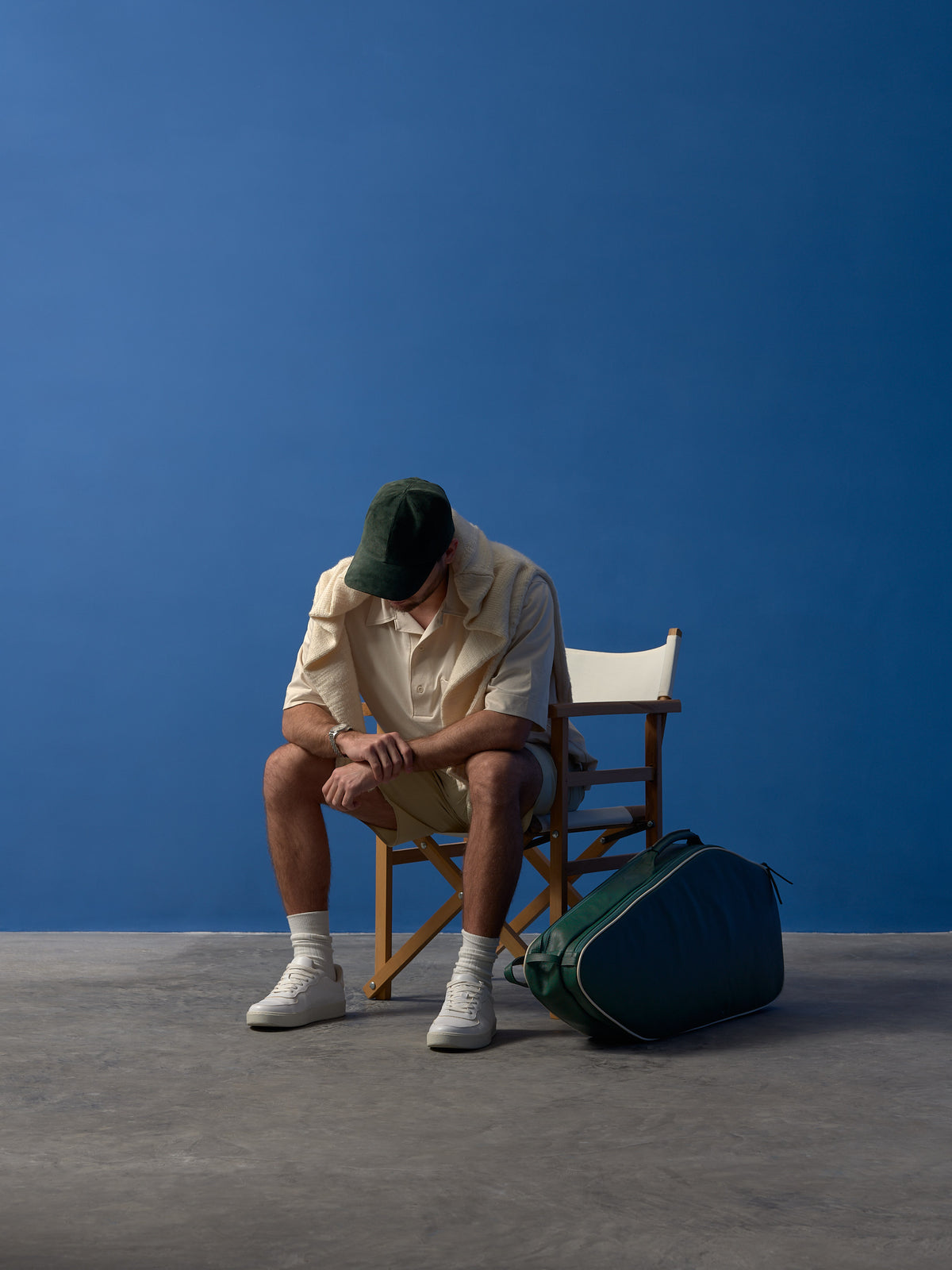 A moody shot of a man, wearing a green suede cap and a beige sweater draped over his shoulders, sitting in a wooden director's chair with his head bowed, the green leather tennis bag on the floor next to him.