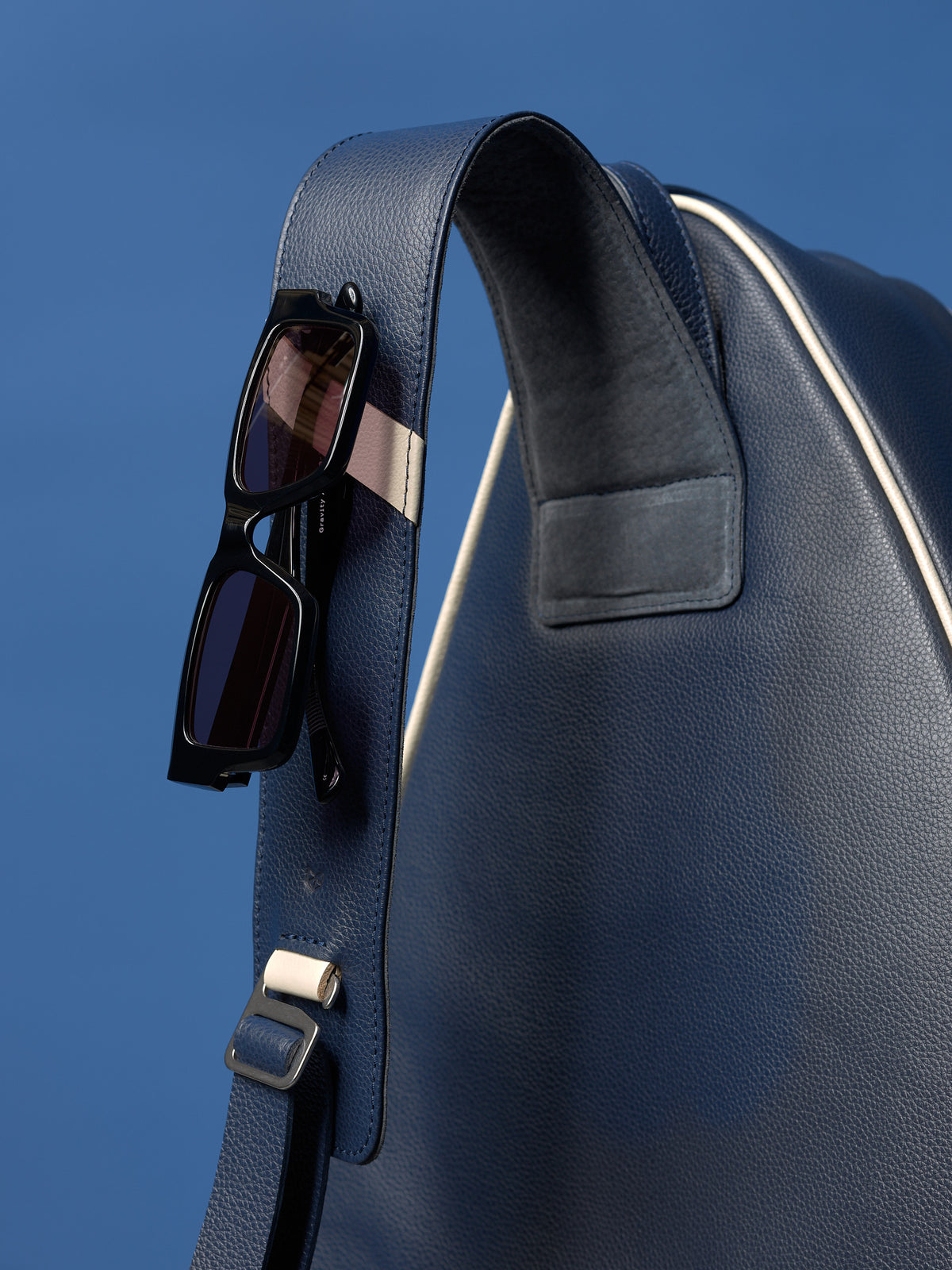 Close-up of black sunglasses clipped to the navy blue leather shoulder strap of the tennis bag.