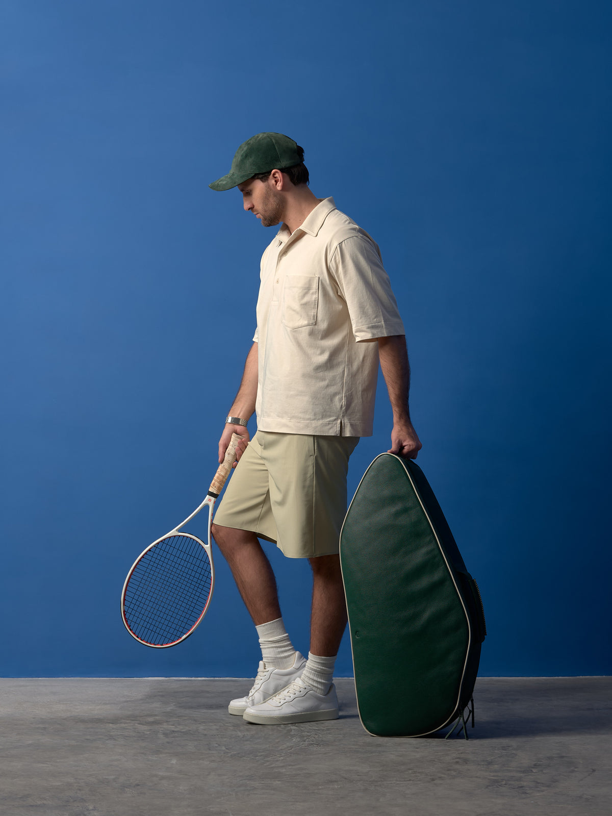 Man in cream polo and green cap holding a forest green leather tennis bag with a white racket.