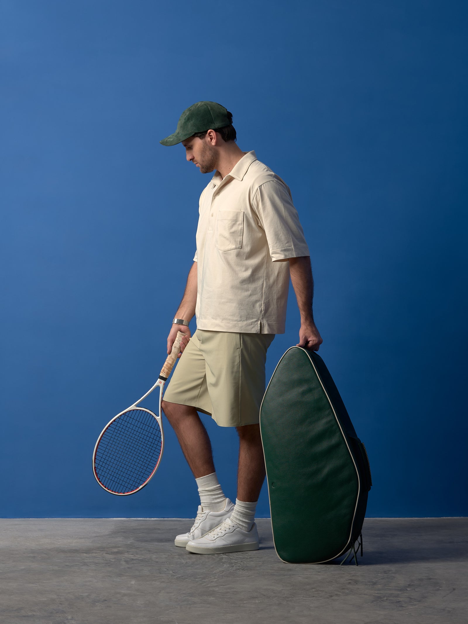 Man in cream polo and green cap holding a forest green leather tennis bag with a white racket.
