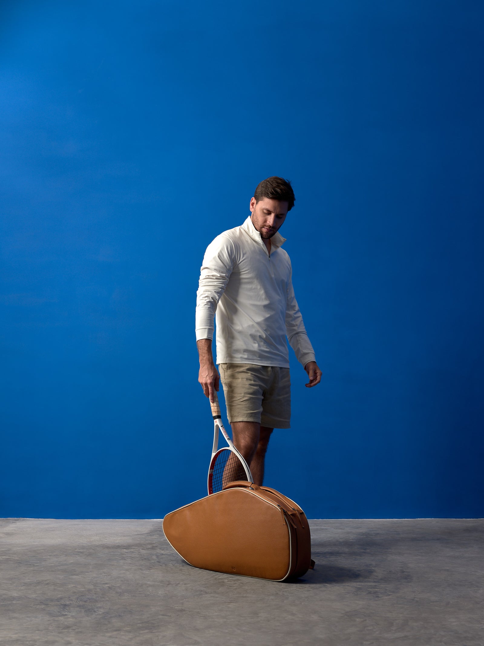 Man standing with a tan full-grain leather tennis bag and racket against a vibrant blue studio backdrop.