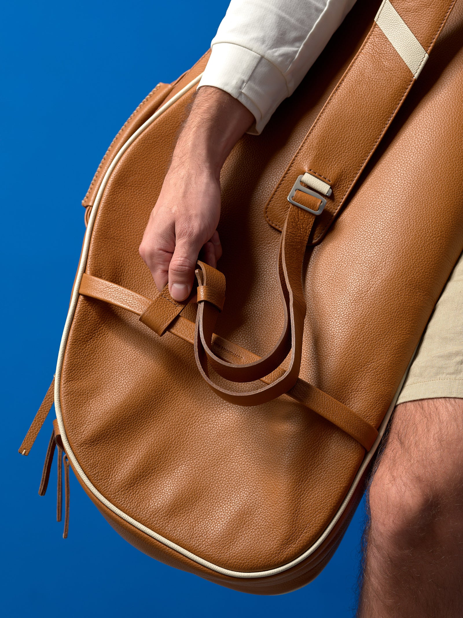 Detailed close-up of a hand securing a loose strap on a designer tan leather tennis bag against a blue background.
