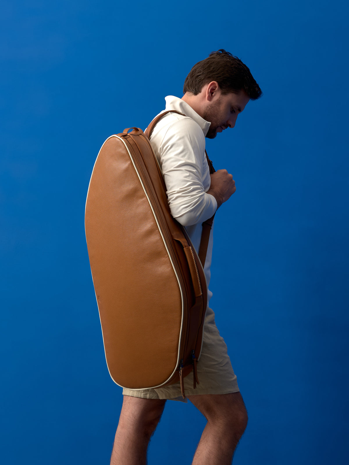Profile view of a man carrying a large tan leather tennis racket bag over his shoulder.