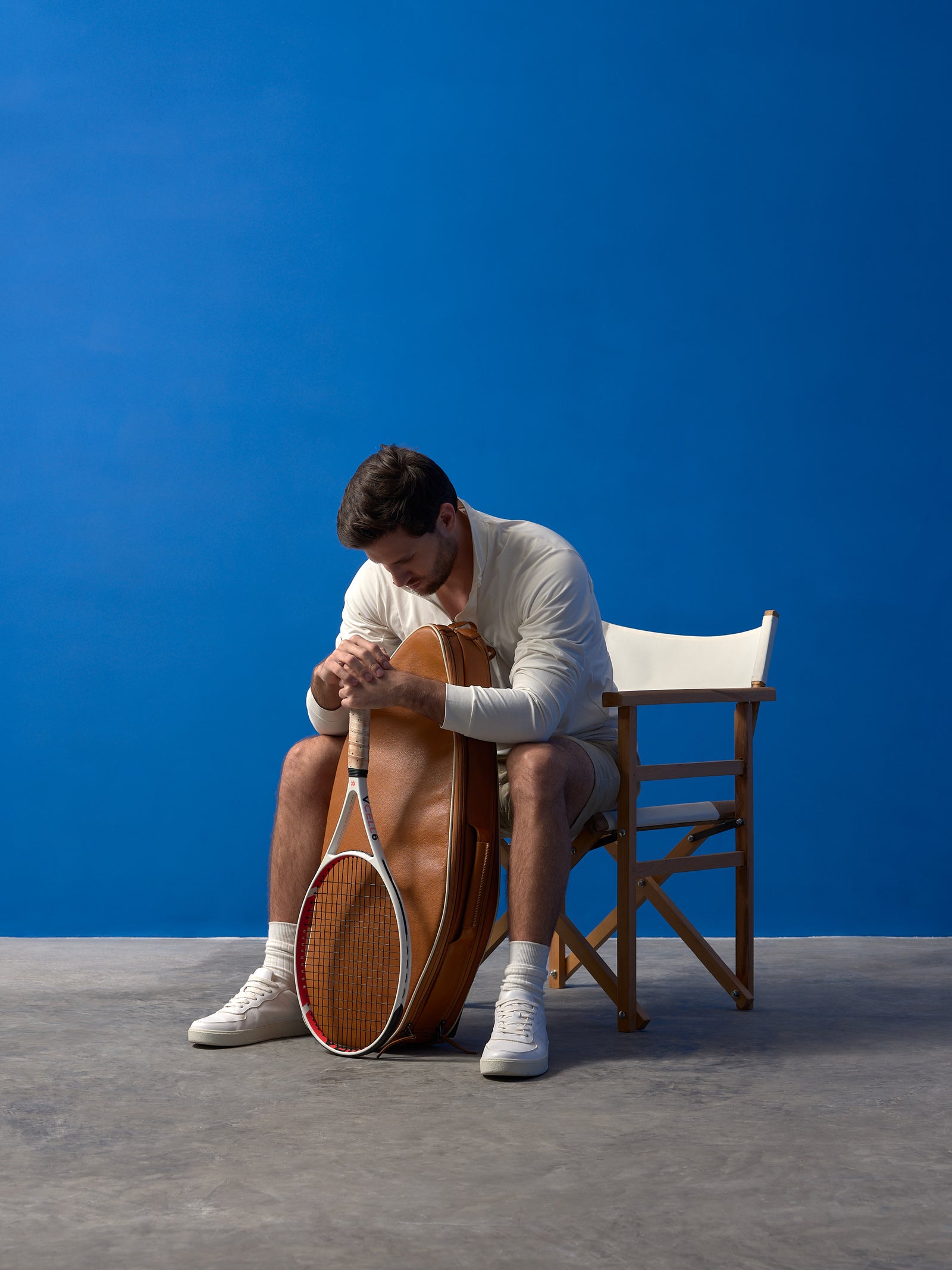 Man sitting in a wooden director's chair leaning on a tan leather tennis bag with a racket.