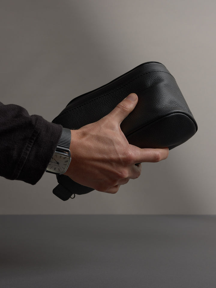 Close-up of a man's hand holding the black full-grain leather Capra dopp kit, showing texture.