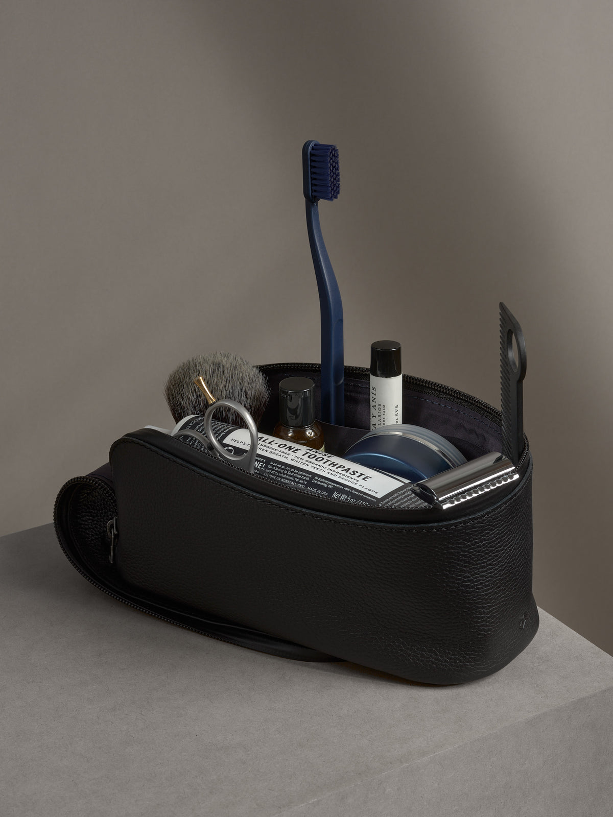 Open black leather toiletry bag filled with grooming essentials: toothpaste, toothbrush, razor, and brush.