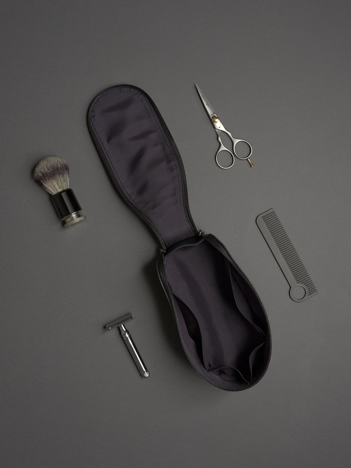 Capra Leather black dopp kit open, featuring razor, shaving brush, scissors, and comb on a gray background.