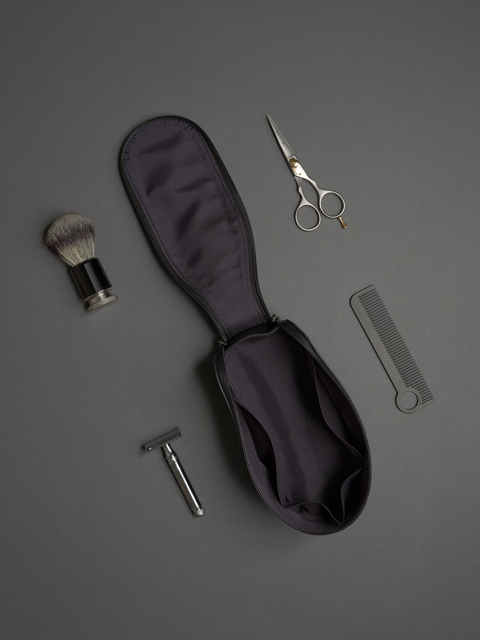 Capra Leather black dopp kit open, featuring razor, shaving brush, scissors, and comb on a gray background.