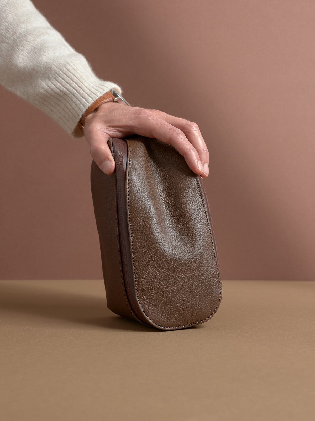 A hand resting on the closed, upright brown Capra Leather Scout Toiletry Bag.