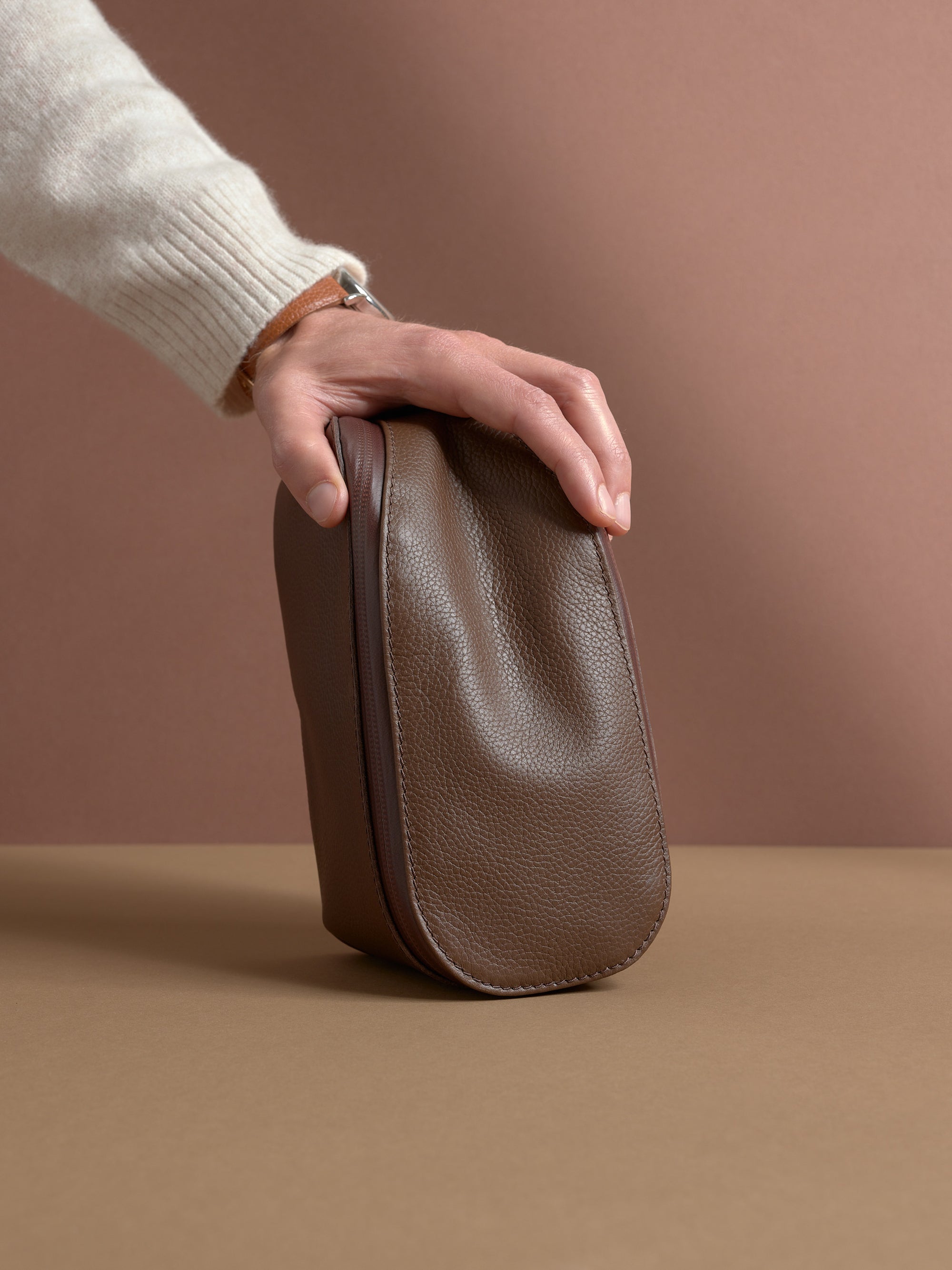 A hand resting on the closed, upright brown Capra Leather Scout Toiletry Bag.