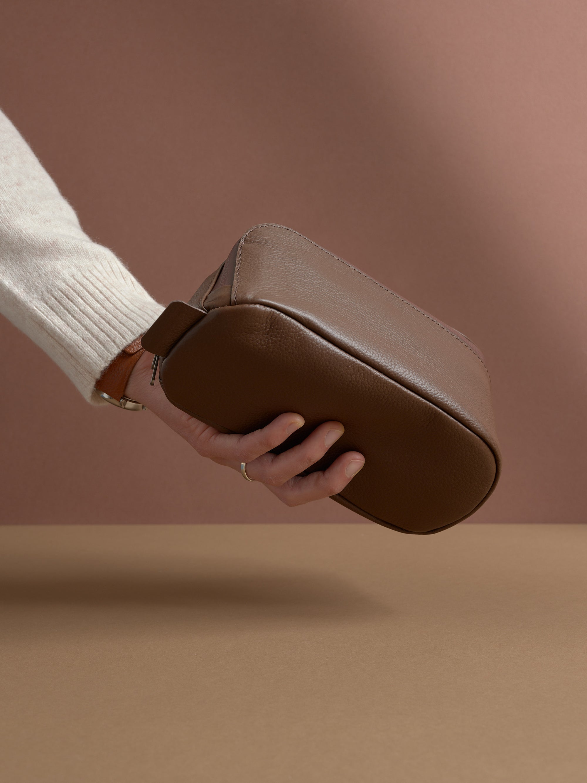 A hand holding the closed brown Capra Leather Toiletry Bag by the side profile.