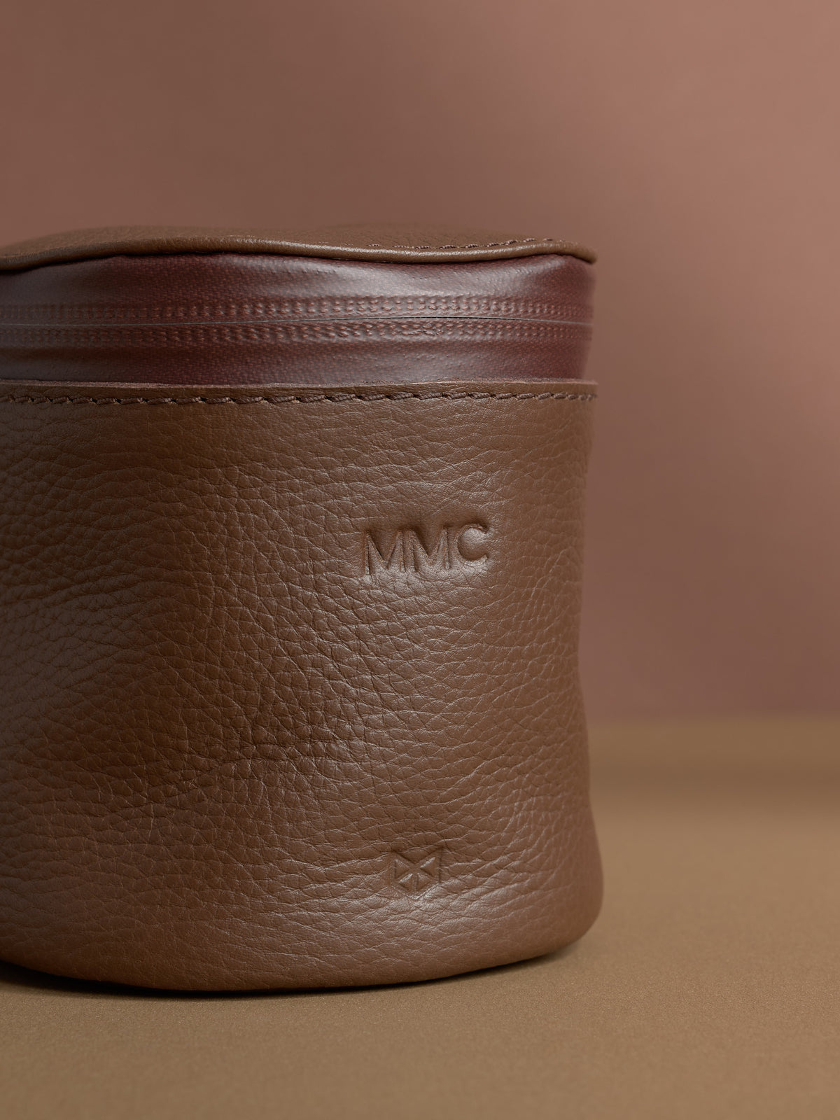 Close-up of a personalized brown Capra leather cylindrical toiletry bag. The leather has a pebbled texture, and the initials "MMC" are debossed near the top