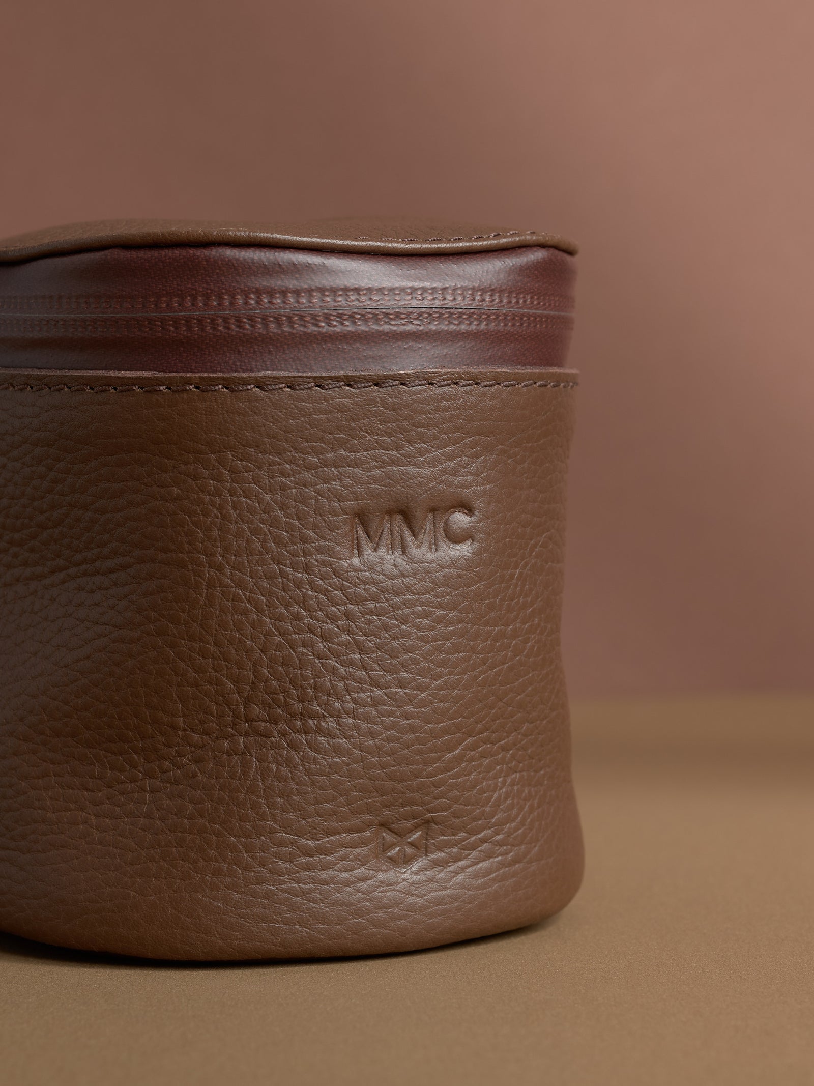 Close-up of a personalized brown Capra leather cylindrical toiletry bag. The leather has a pebbled texture, and the initials "MMC" are debossed near the top