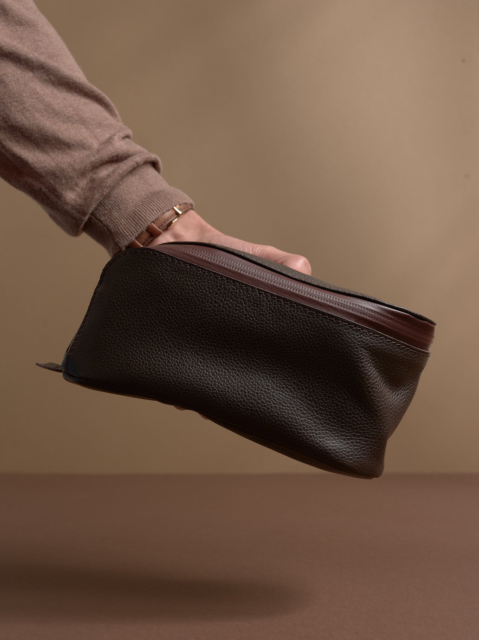 A hand holding the closed dark brown Capra Leather Toiletry Bag by the side.