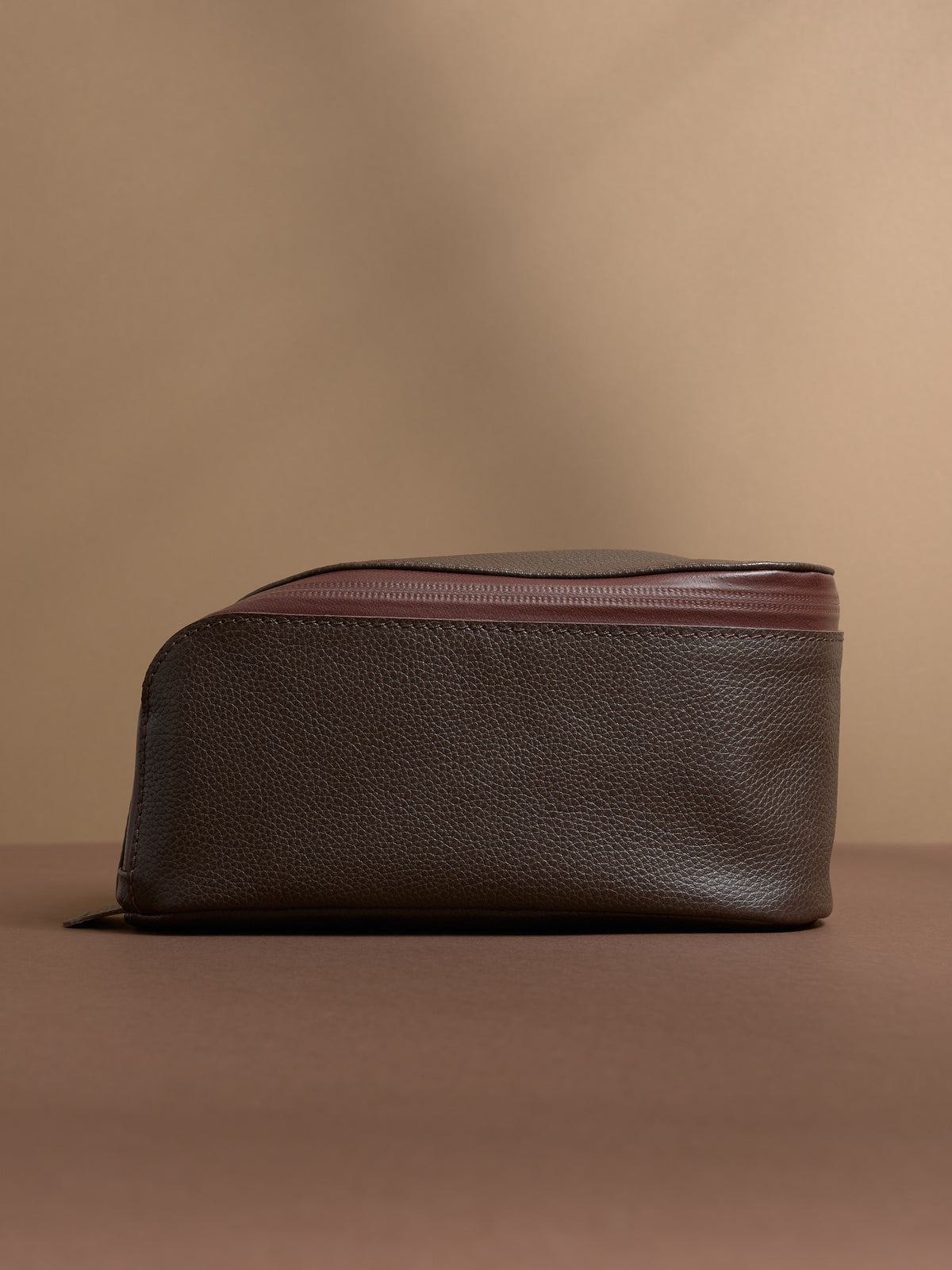 Side profile of the closed dark brown Capra Leather Toiletry Bag, showing its curved top edge.
