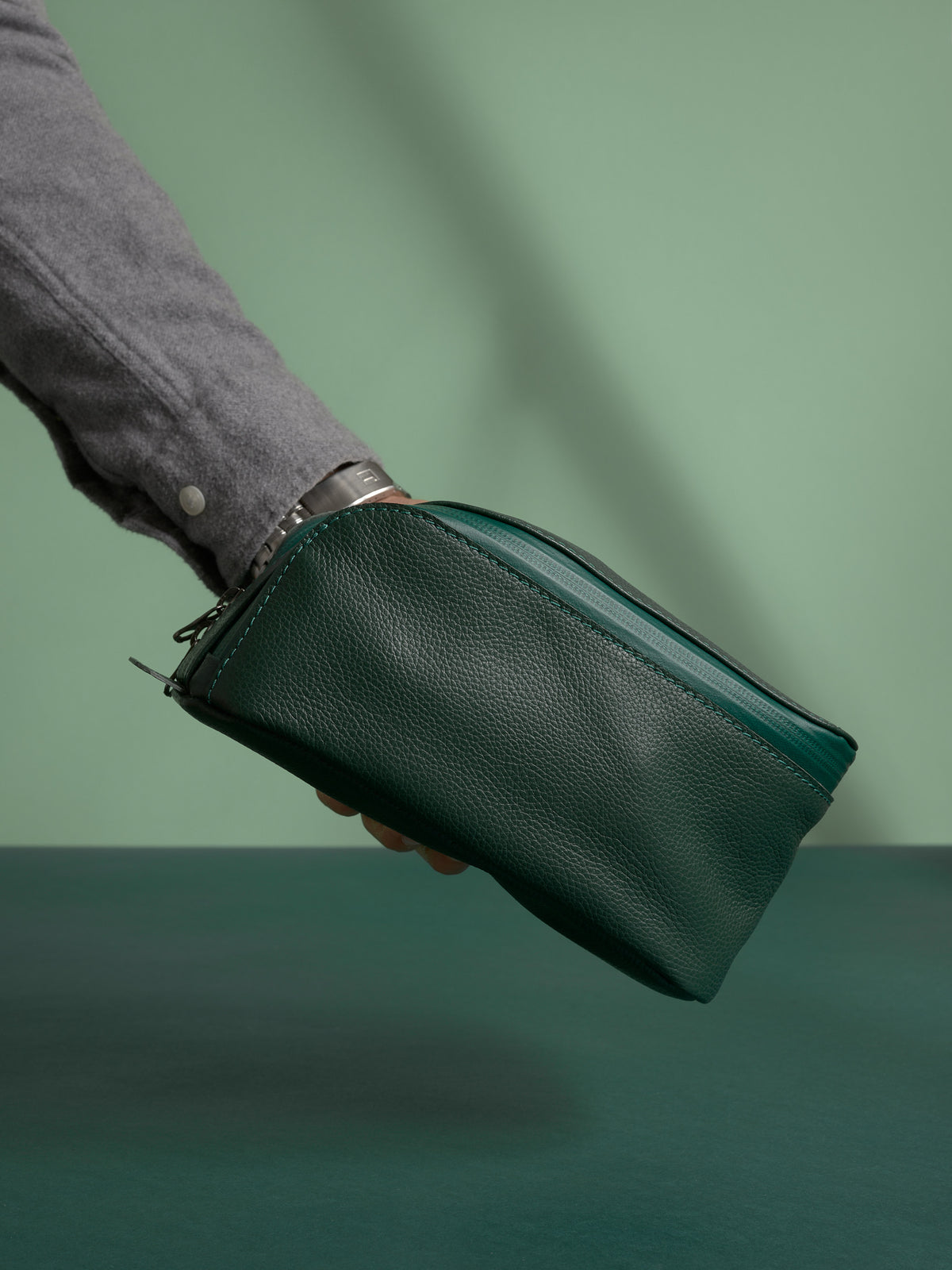 A hand holding the closed, deep green Capra Leather Toiletry Bag by the side loop.