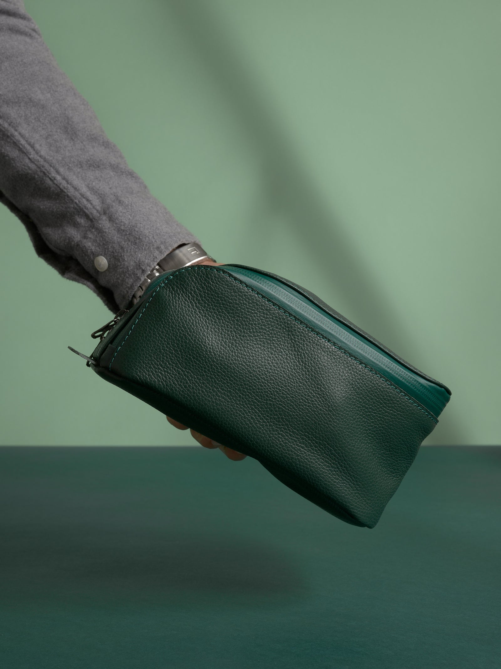 A hand holding the closed, deep green Capra Leather Toiletry Bag by the side loop.