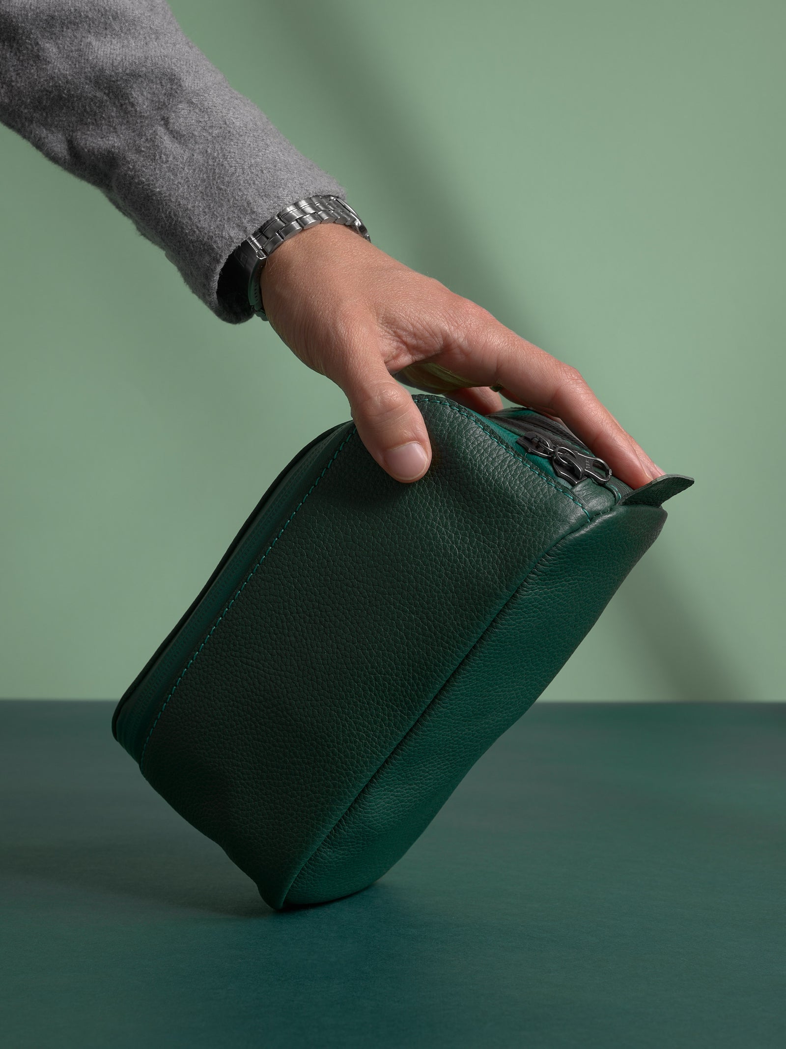 Hand holding the dark green Capra Leather Toiletry Bag upright, showing the base and stitching.