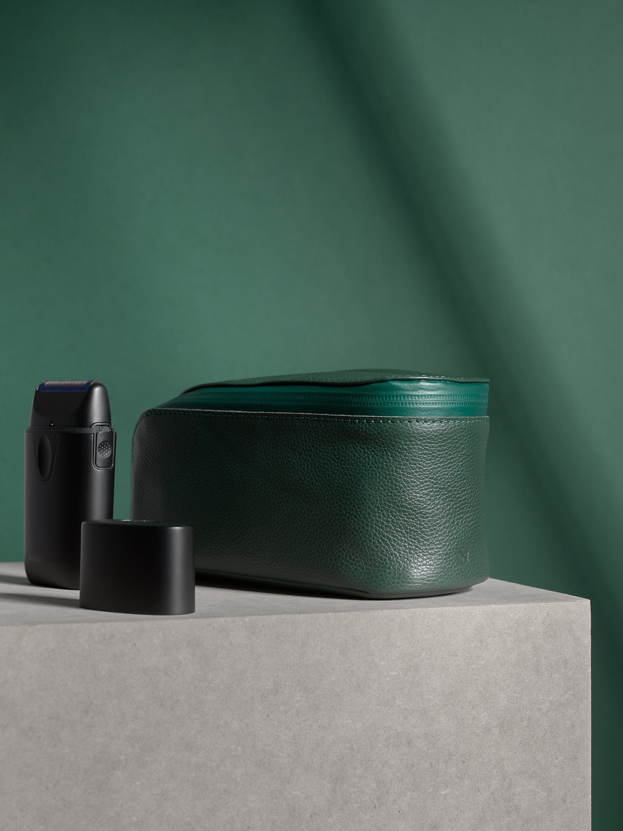 Closed deep green Capra Leather Toiletry Bag standing next to an electric shaver.