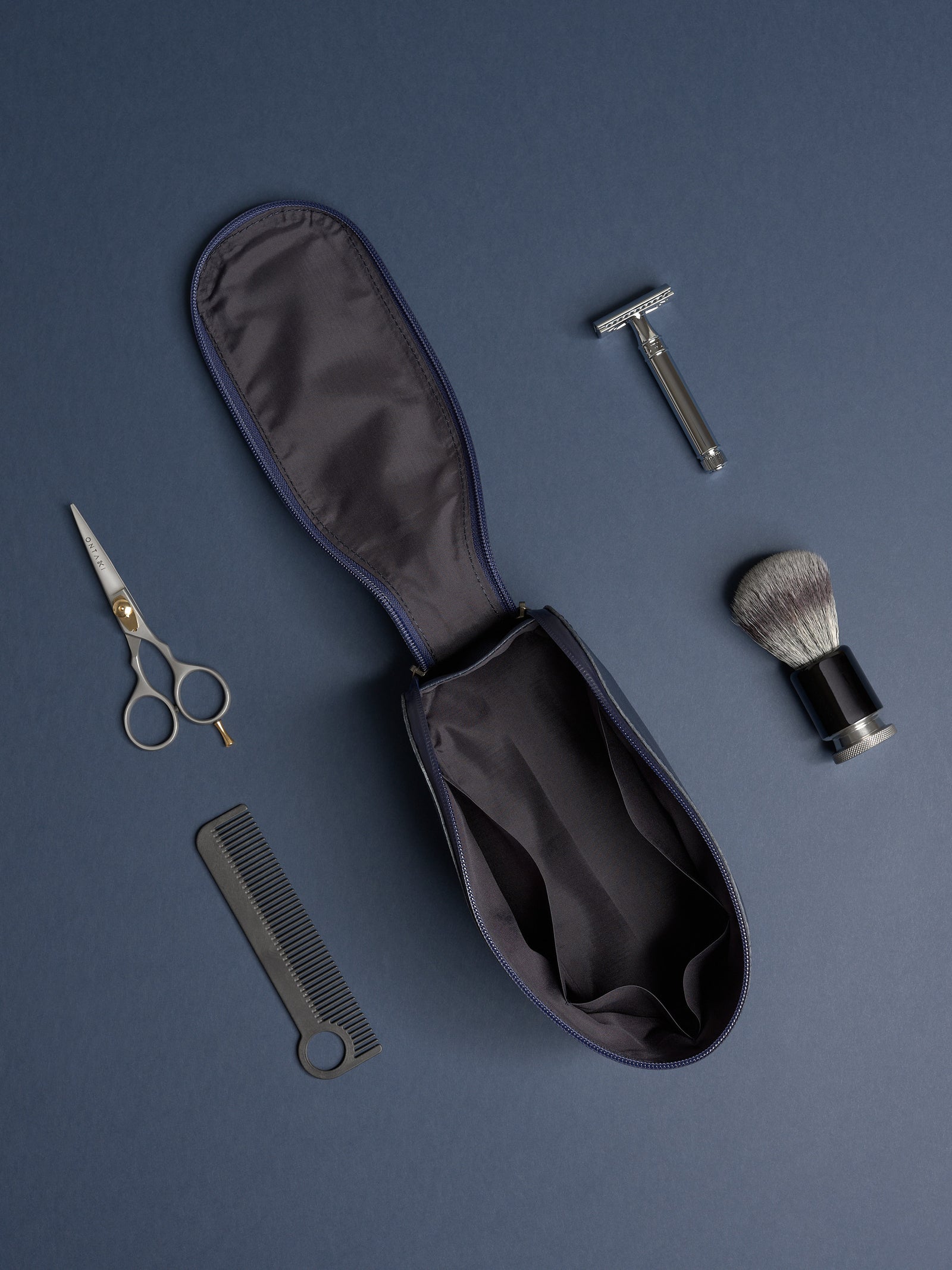 Empty, open navy Capra leather toiletry bag with grooming tools: safety razor, shaving brush, comb, and scissors.