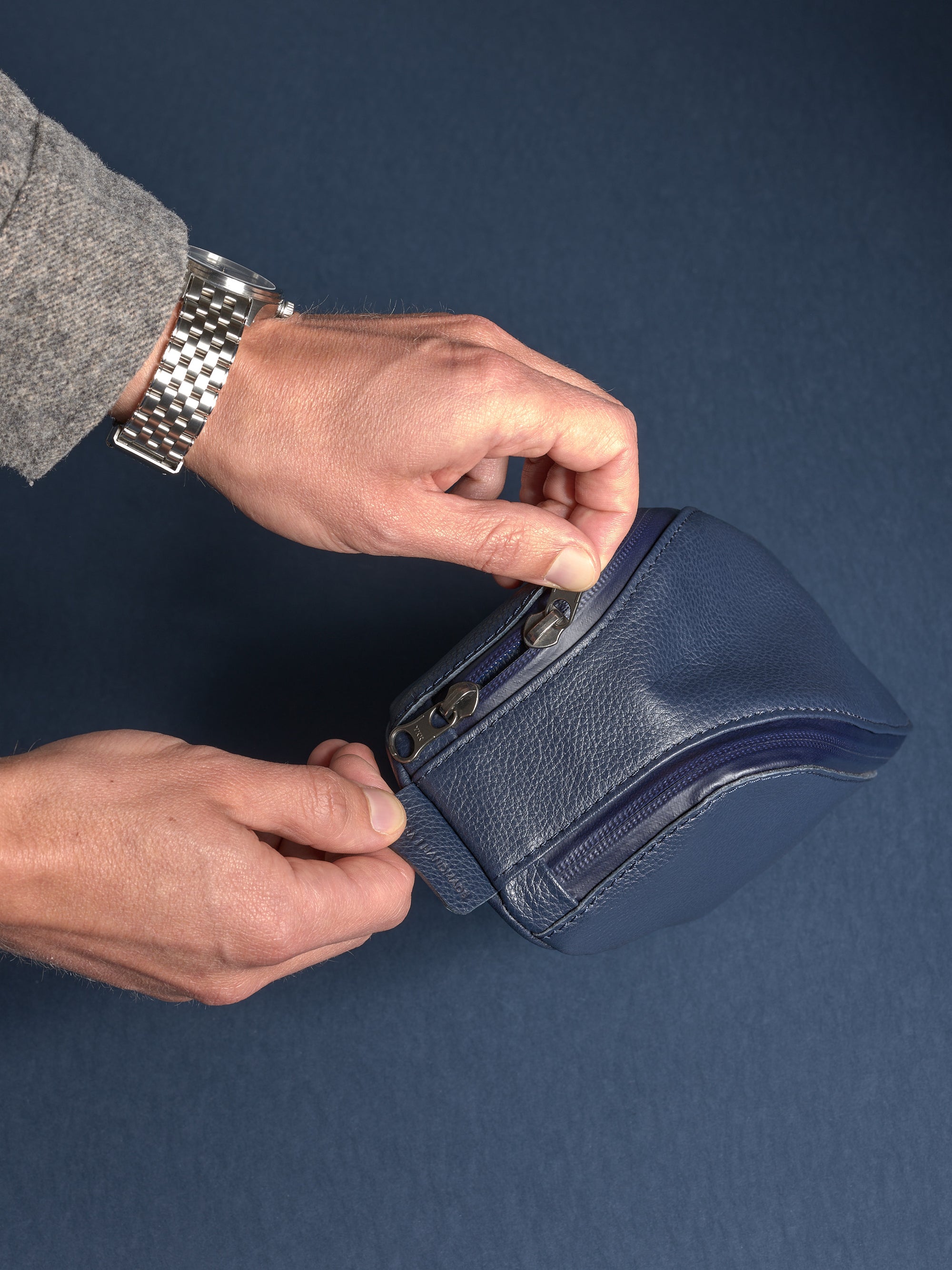 Man's hands opening the navy Capra leather dopp kit's zipper, showing the quality hardware and rich texture.