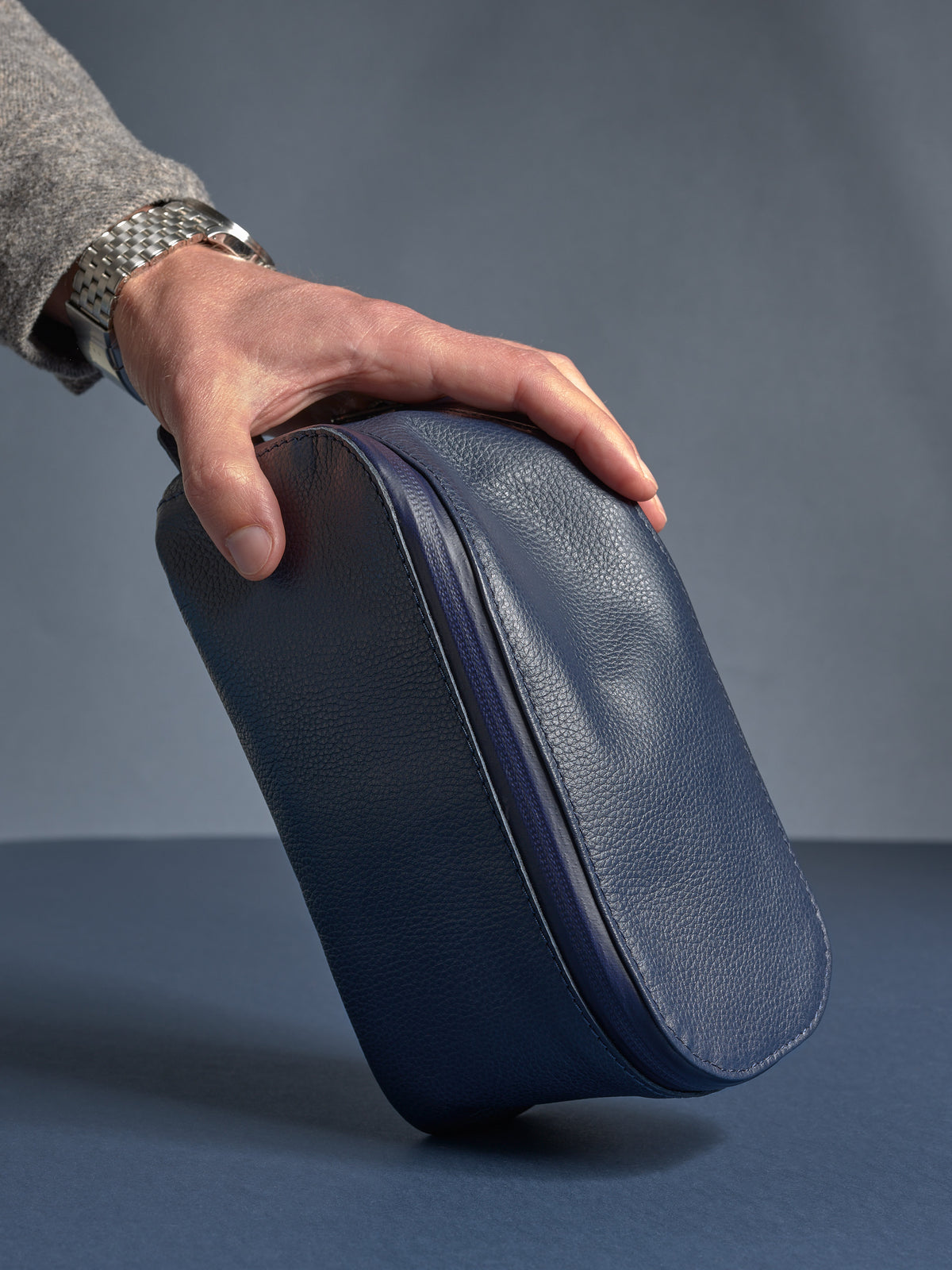 Man holding the navy Capra leather Scout Toiletry Bag by its handle, highlighting its compact, travel-ready shape.