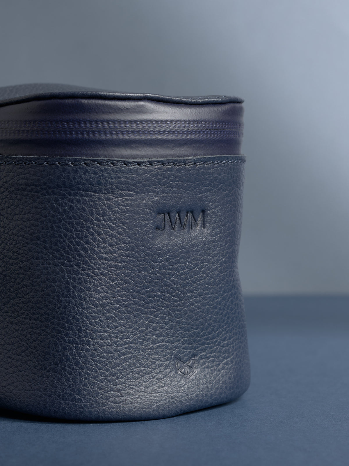 Close-up of a personalized navy blue Capra leather cylindrical toiletry bag. The leather has a pebbled texture, and the initials "JWM" are debossed near the top.