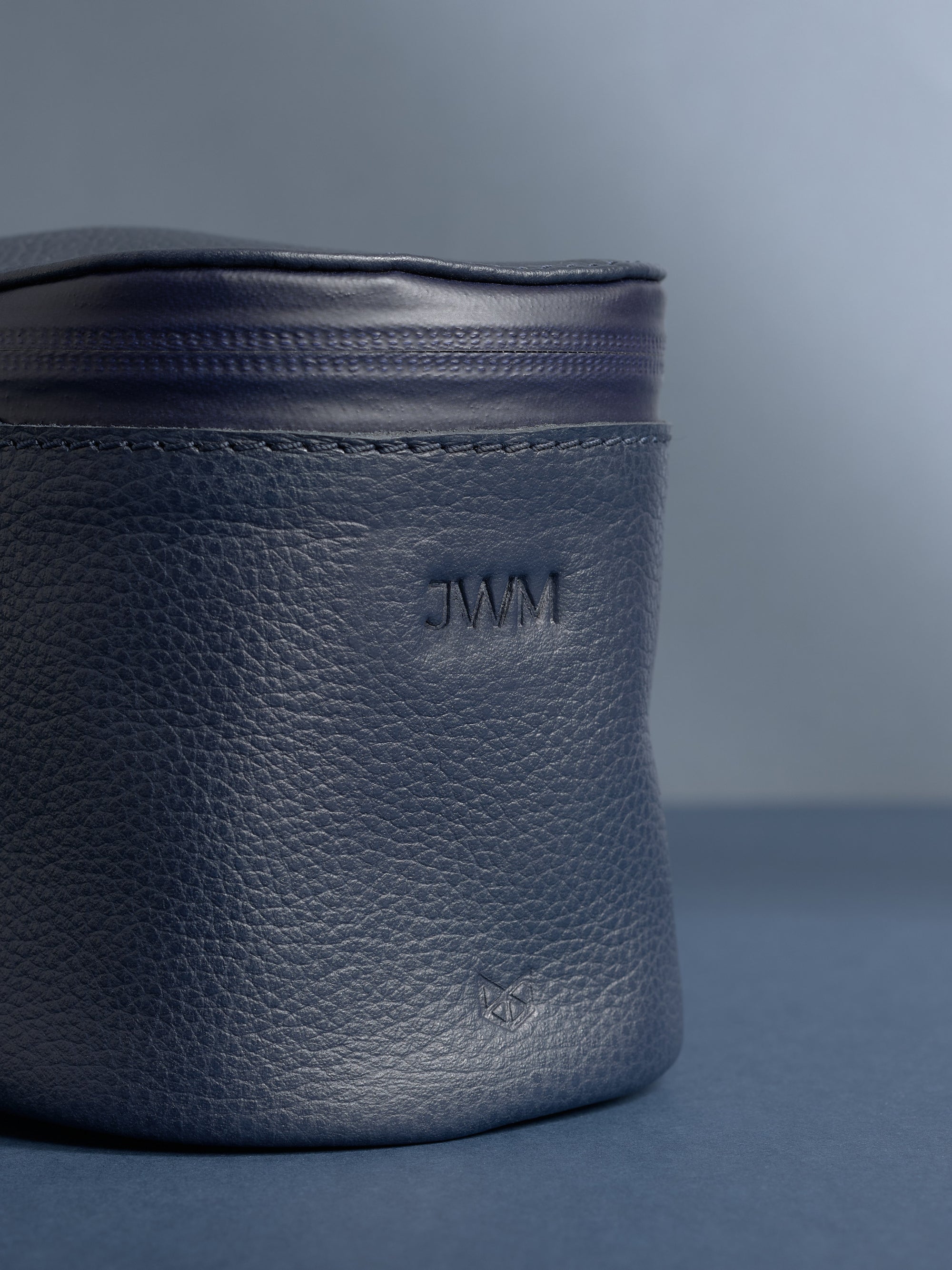 Close-up of a personalized navy blue Capra leather cylindrical toiletry bag. The leather has a pebbled texture, and the initials "JWM" are debossed near the top.
