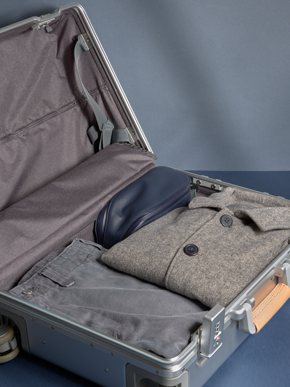 Navy leather toiletry bag packed neatly next to a gray coat and pants inside a silver travel suitcase.