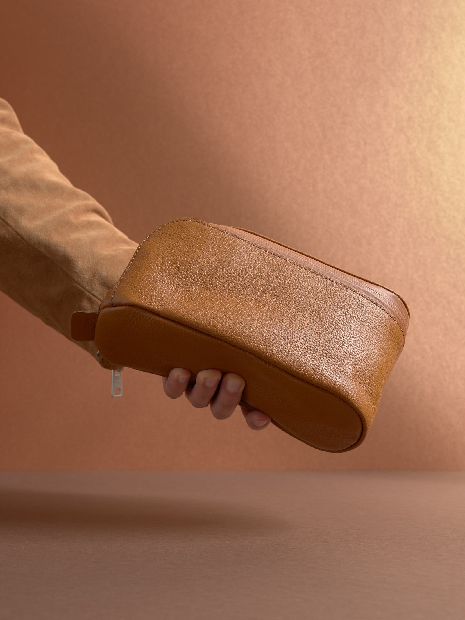 A hand holding the closed tan Capra Leather Toiletry Bag by the side loop, showcasing the side profile.