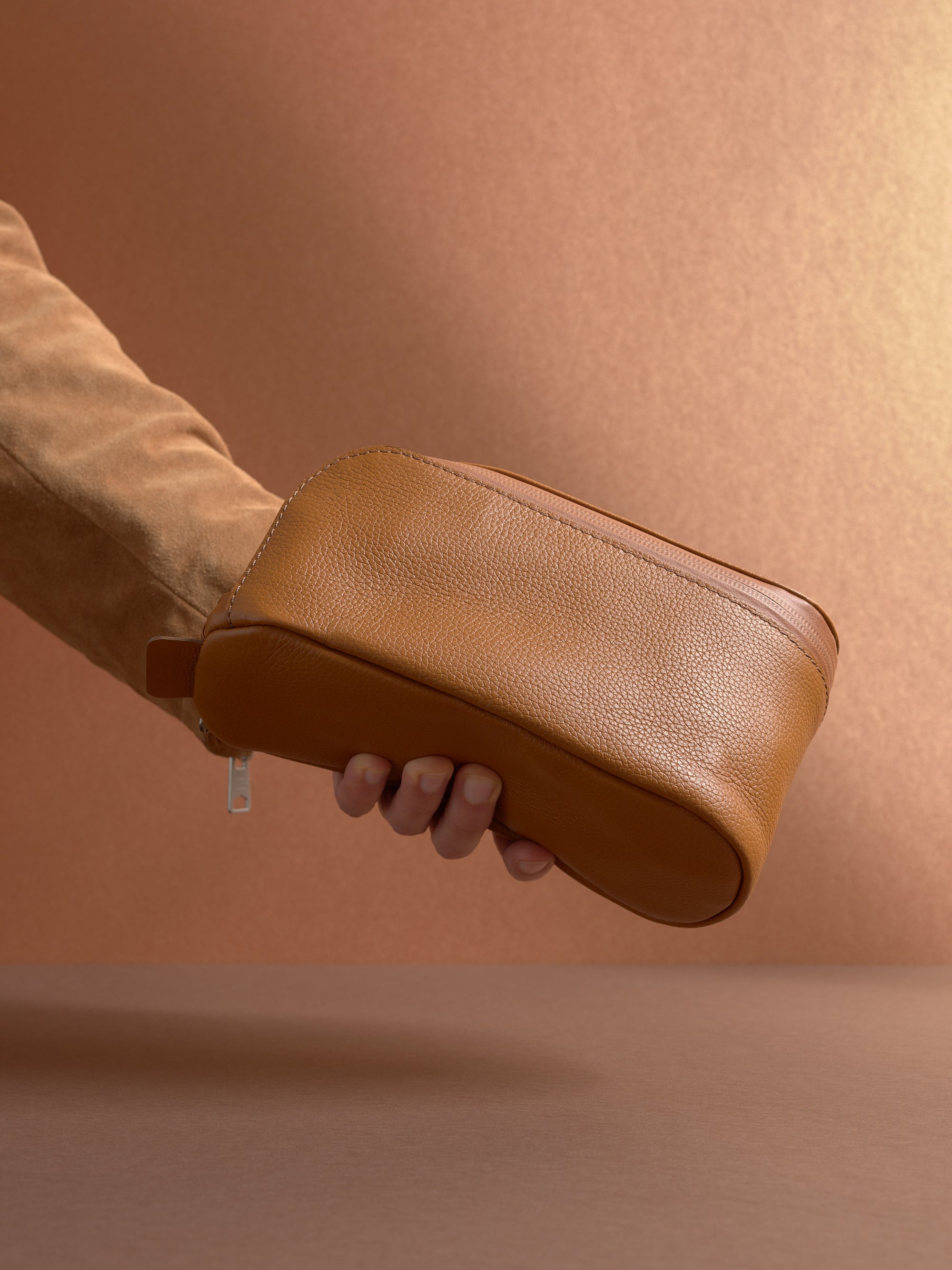 A hand holding the closed tan Capra Leather Toiletry Bag by the side loop, showcasing the side profile.