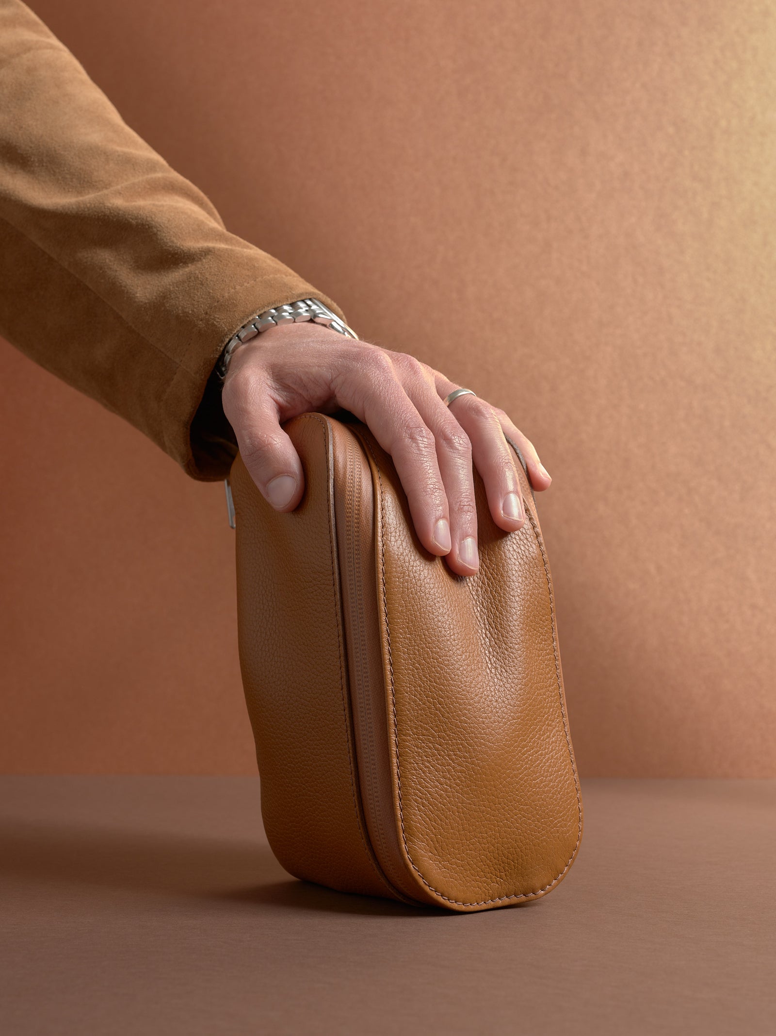 Hand resting on the closed, upright tan Capra Leather Scout Toiletry Bag, highlighting its shape.