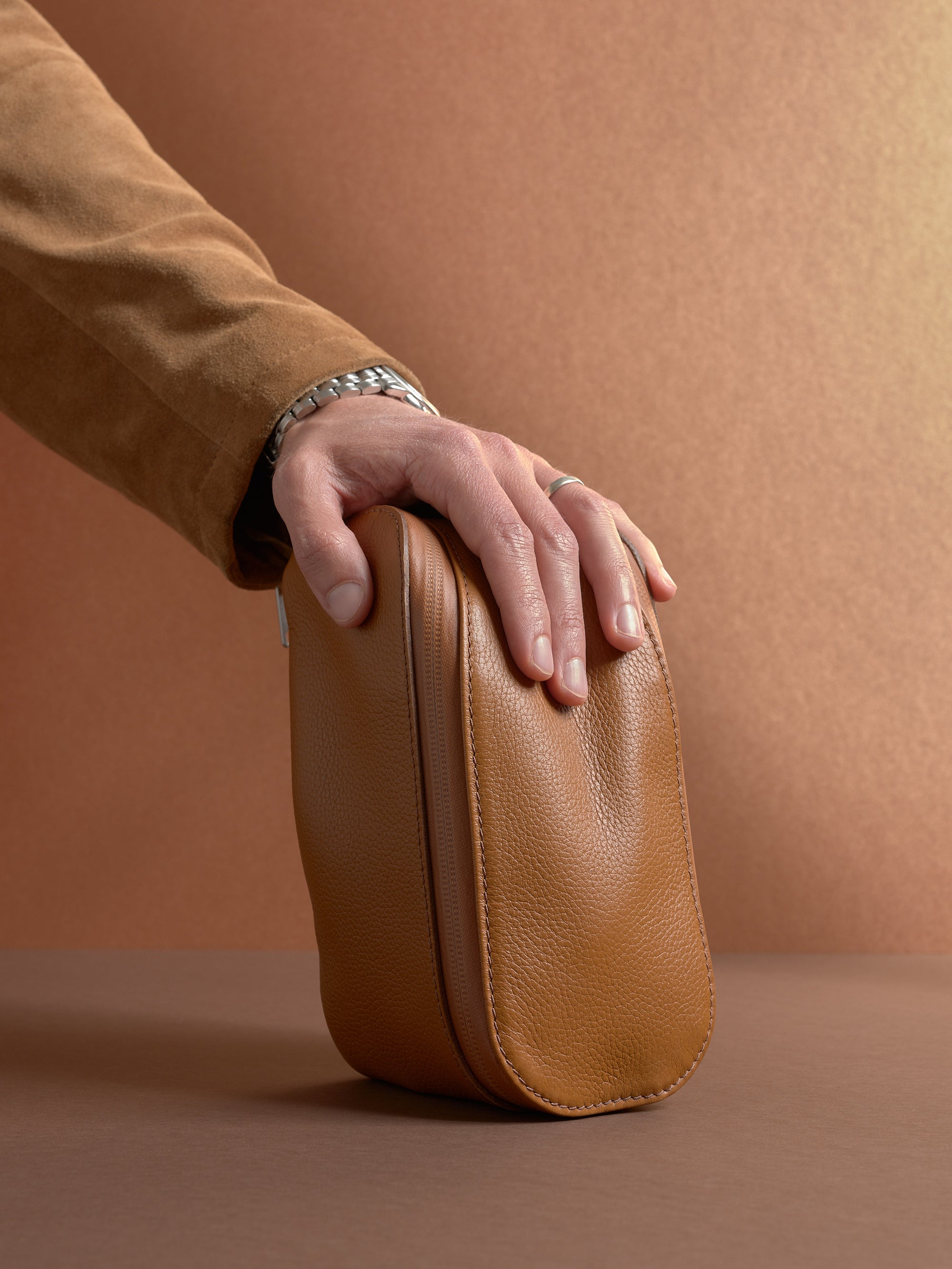 Hand resting on the closed, upright tan Capra Leather Scout Toiletry Bag, highlighting its shape.
