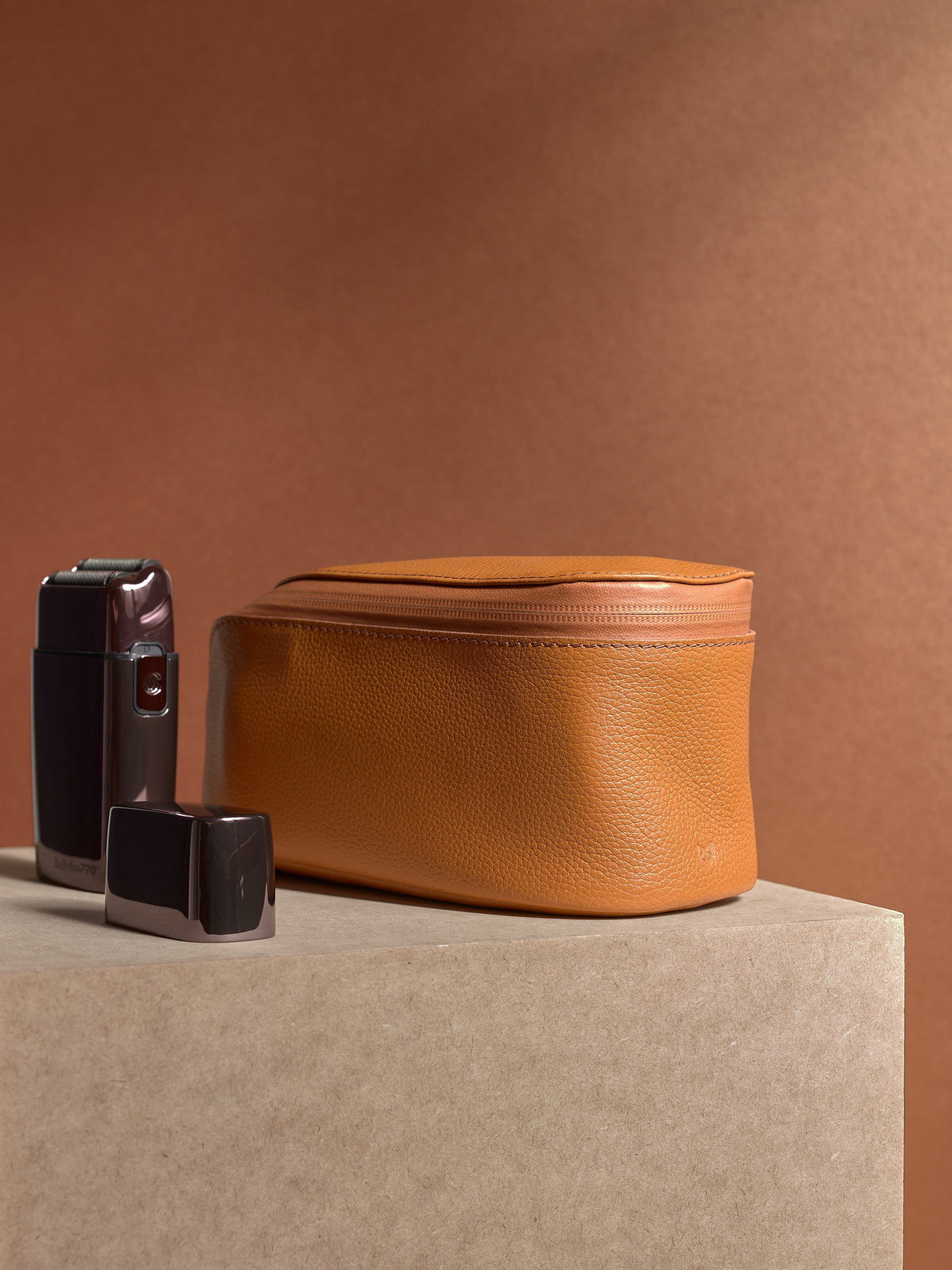 Closed tan Capra Leather Toiletry Bag standing on a podium next to an electric shaver.