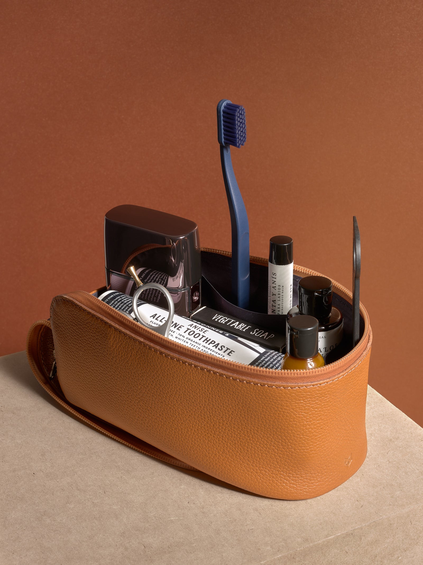 A hand holding the closed tan Capra Leather Toiletry Bag by the side loop, showcasing the side profile.