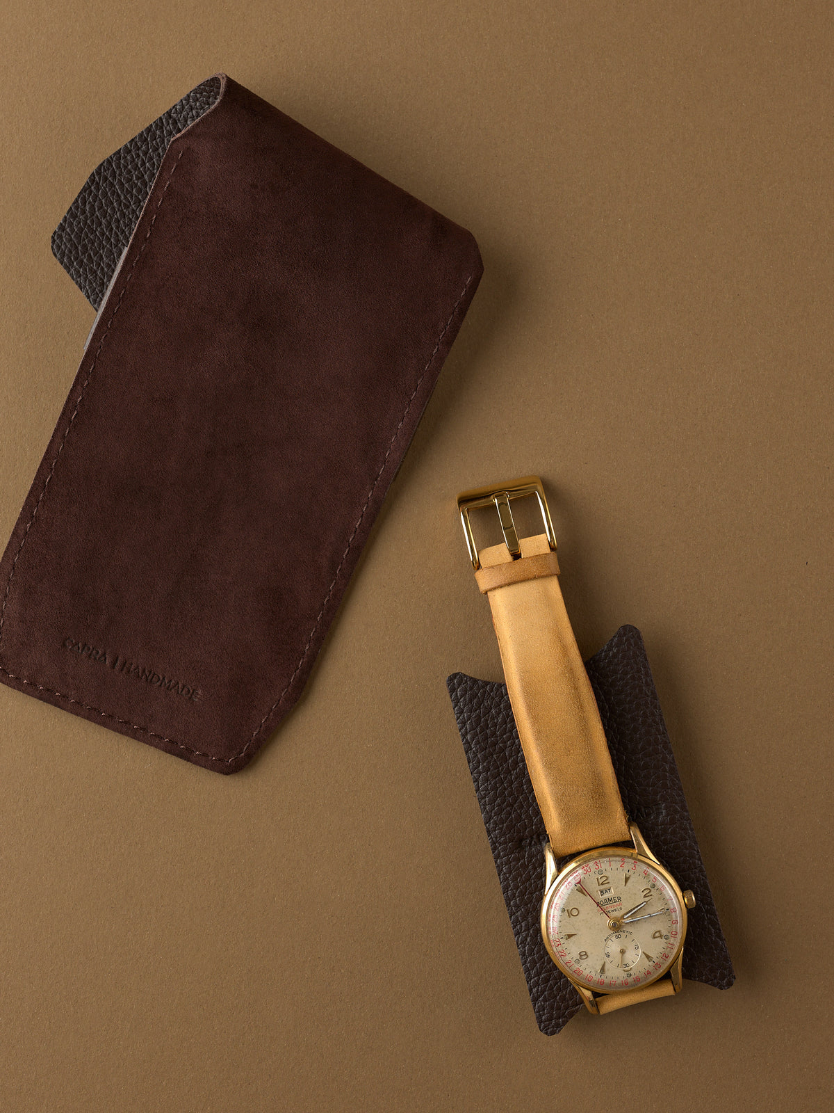 The Capra Handmade brown suede watch pouch shown next to a classic watch resting on its protective pebbled leather insert.