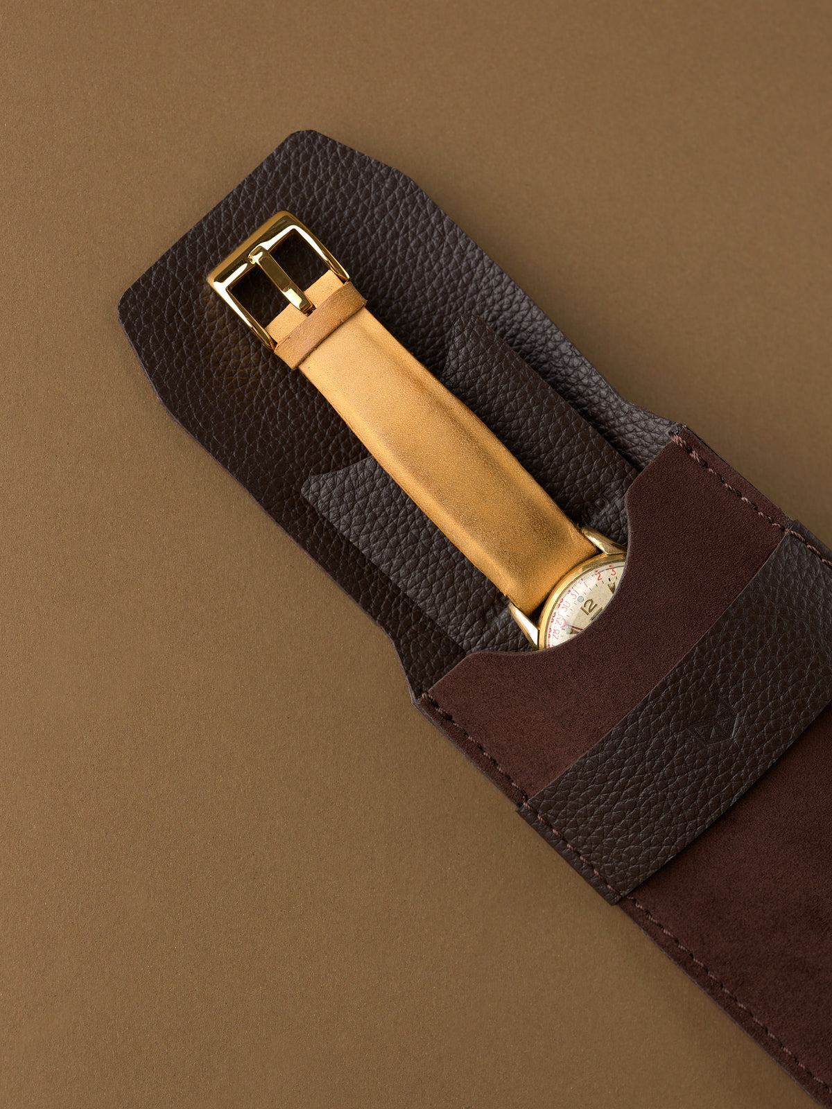 A gold watch with a tan leather strap being placed into the soft interior of a brown suede Capra Leather watch pouch.