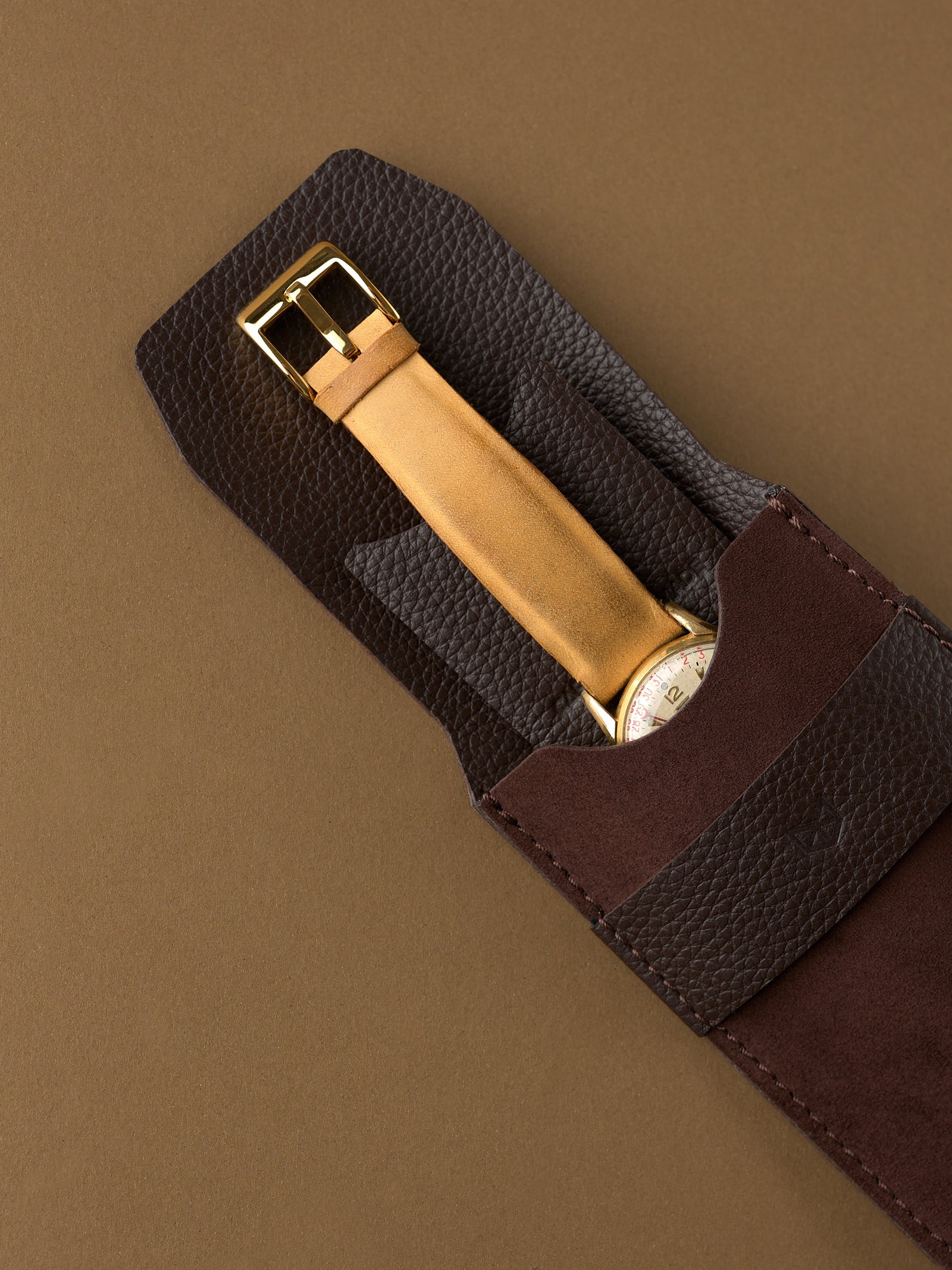 A gold watch with a tan leather strap being placed into the soft interior of a brown suede Capra Leather watch pouch.