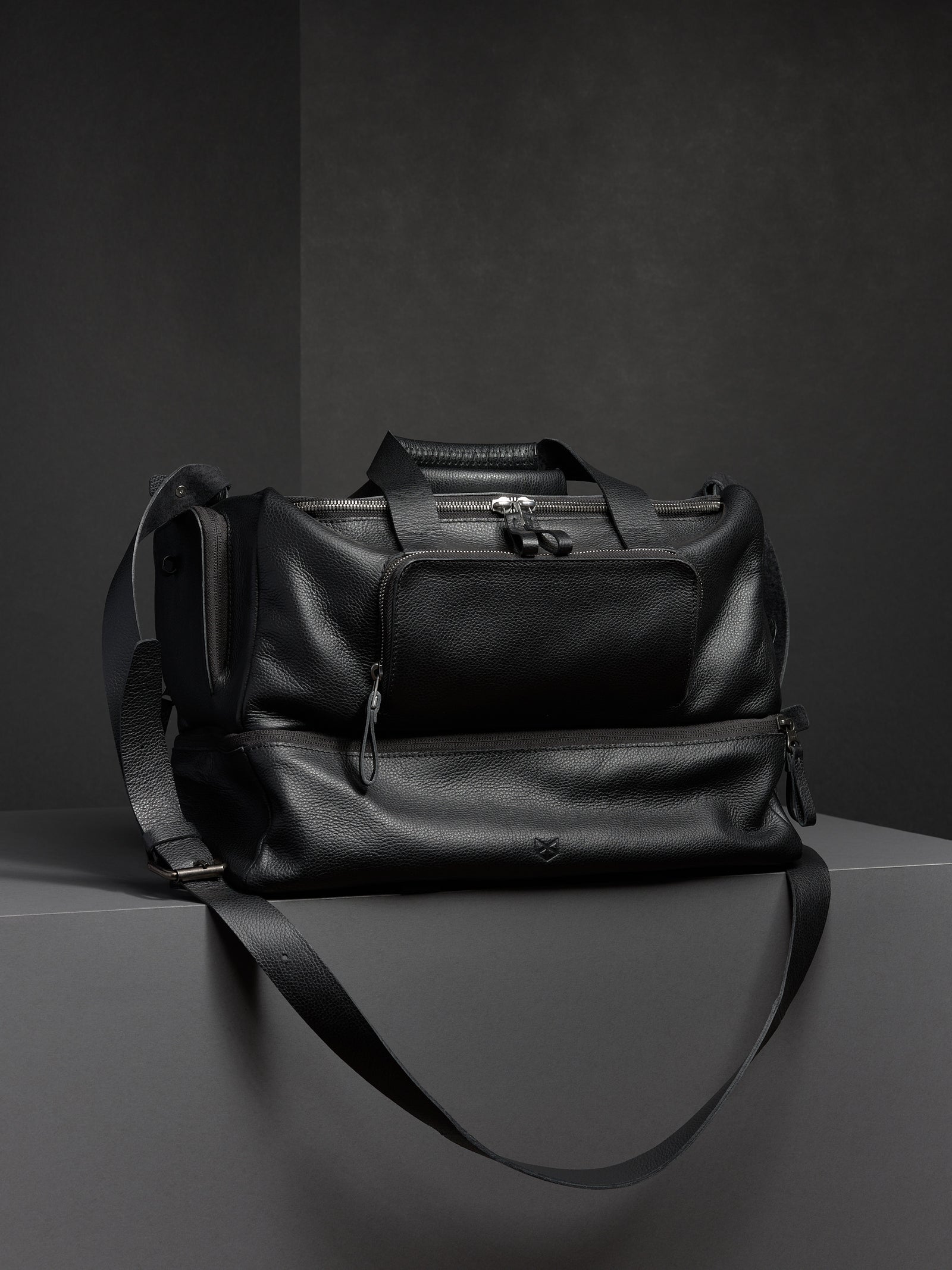 Black leather duffel bag from Capra, showcasing its structured shape and two-part design with a shoulder strap.