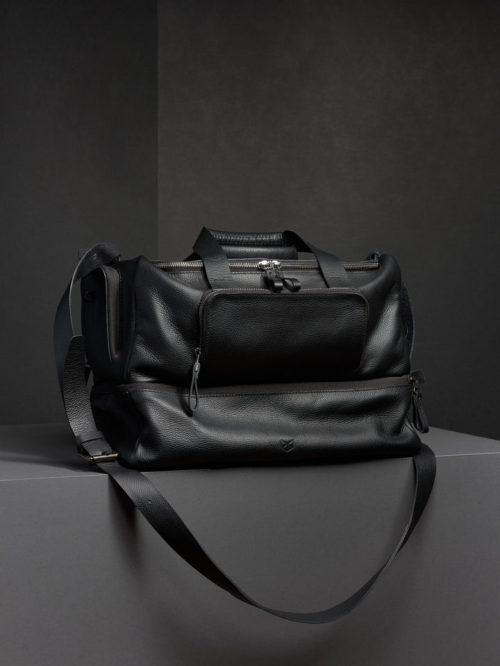 Black leather duffel bag from Capra, showcasing its structured shape and two-part design with a shoulder strap.