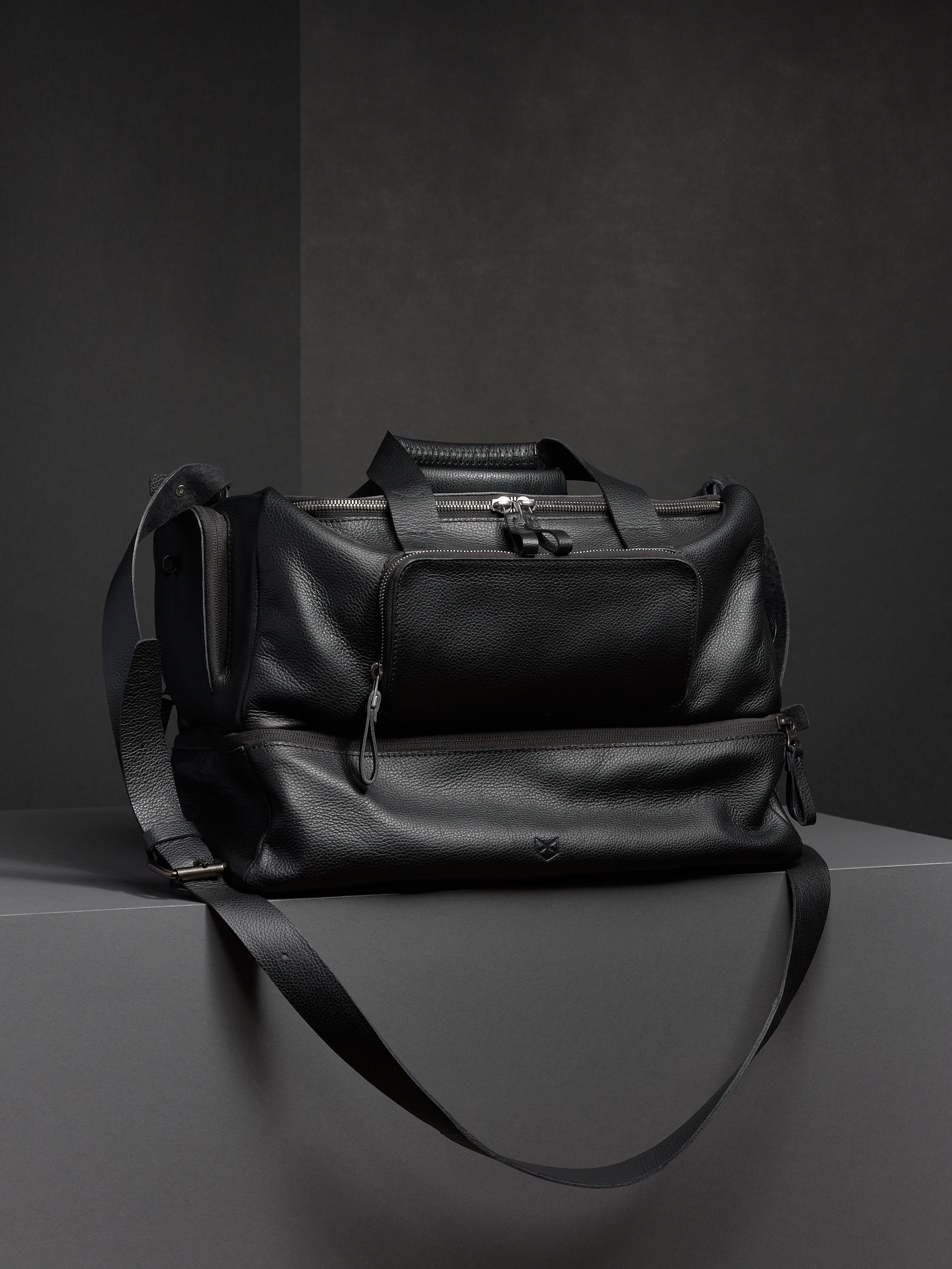Black leather duffel bag from Capra, showcasing its structured shape and two-part design with a shoulder strap.