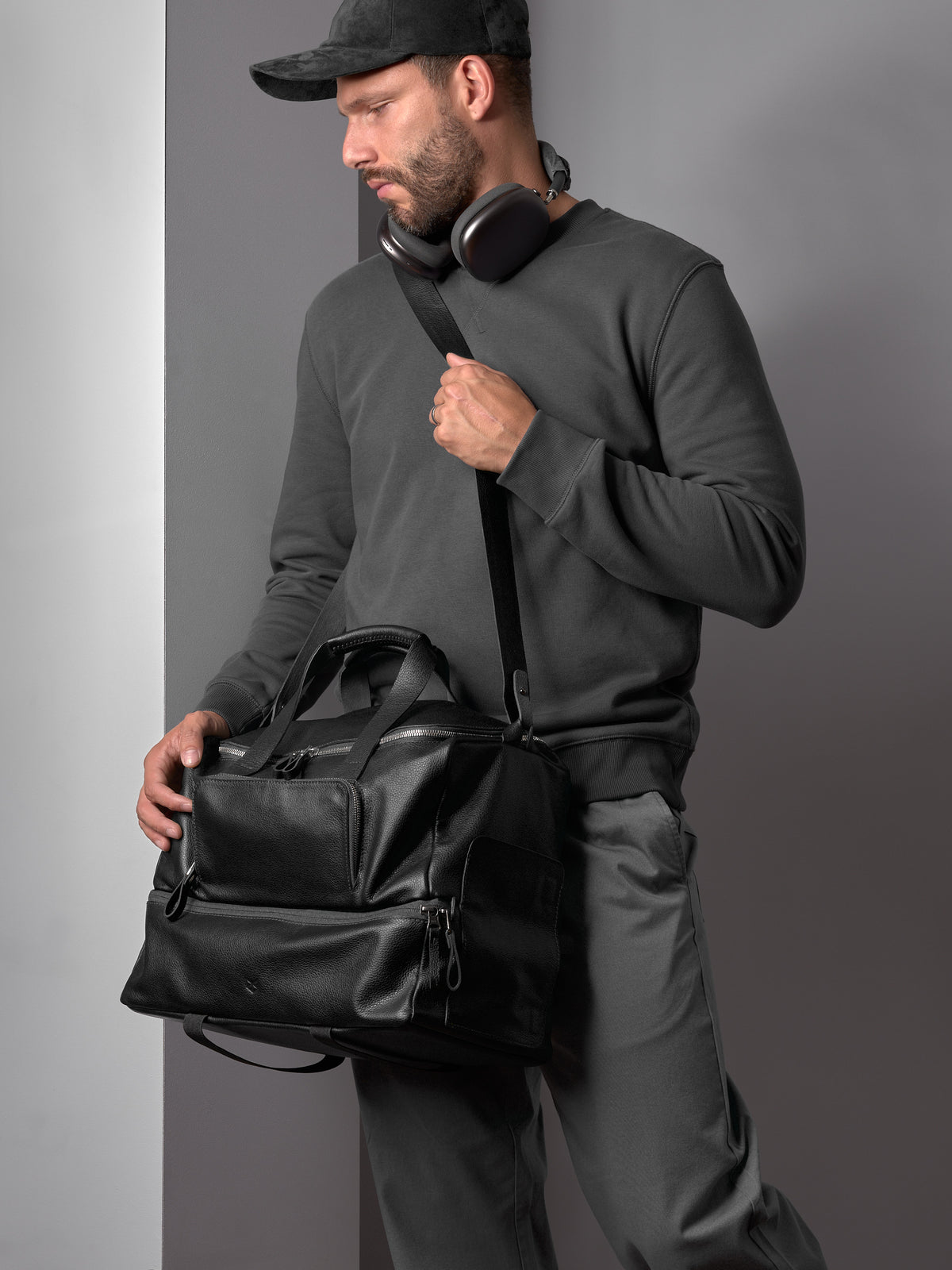 A man wearing a hat and headphones carries the black leather Capra duffel bag, highlighting its versatility.