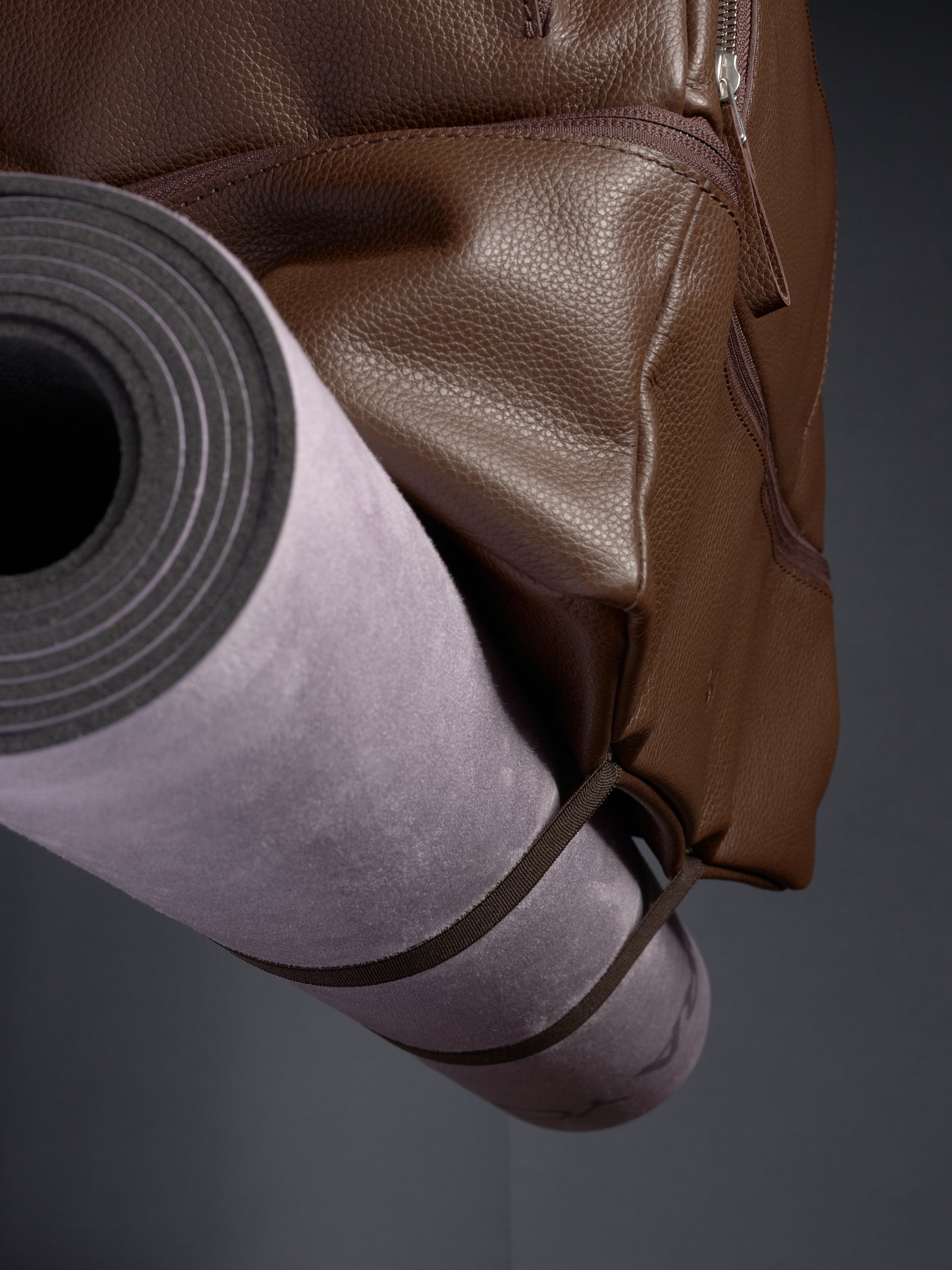 The versatile bottom straps of the brown leather Ventra Holdall securely holding a rolled-up grey yoga mat in place.