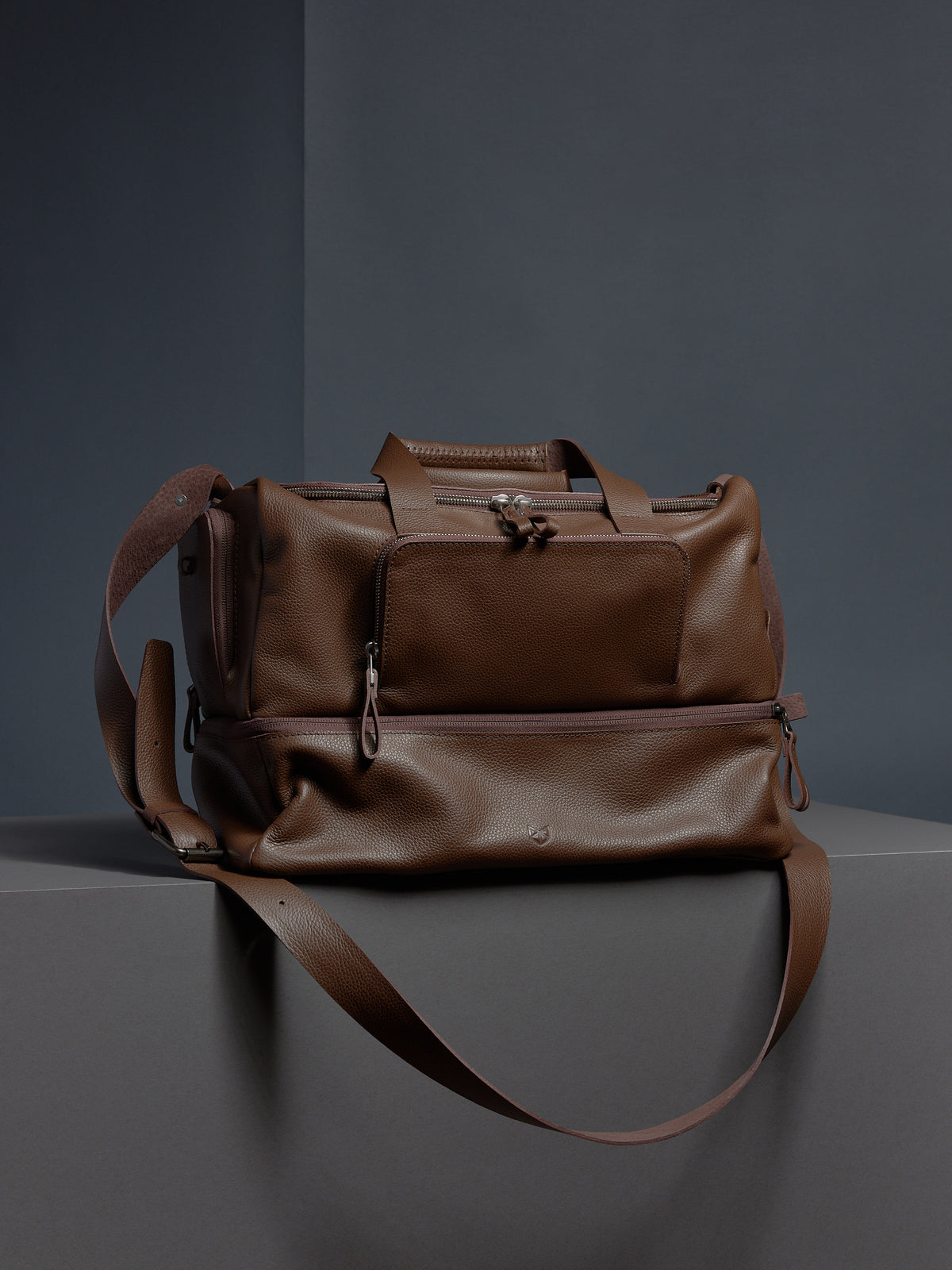 The Ventra Holdall for men in rich brown pebbled leather, shown with its shoulder strap against a grey background.