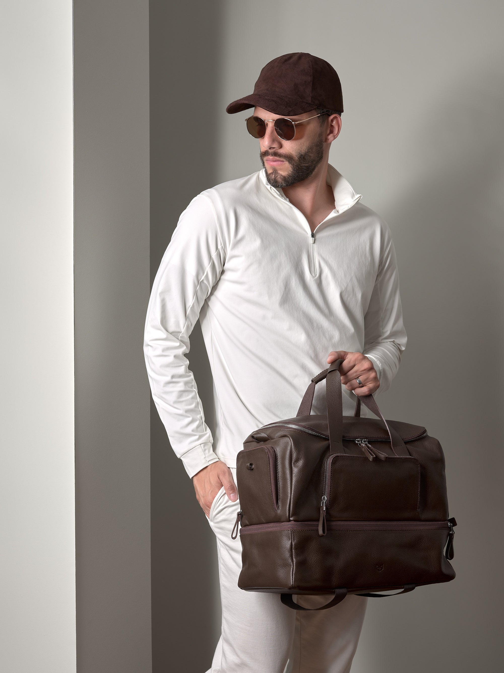 A man with sunglasses and a cap holding the dark brown Capra Leather Ventra Holdall, a luxurious travel bag.