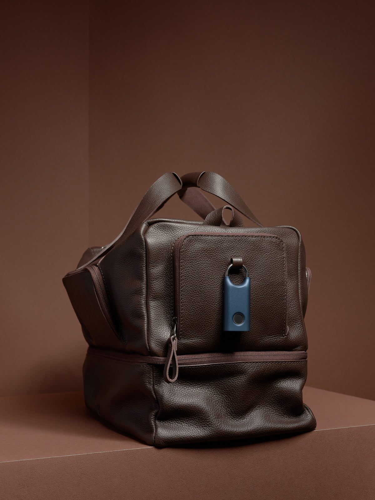 A dark brown leather Ventra Holdall bag on a brown platform with a blue key tracker attached.