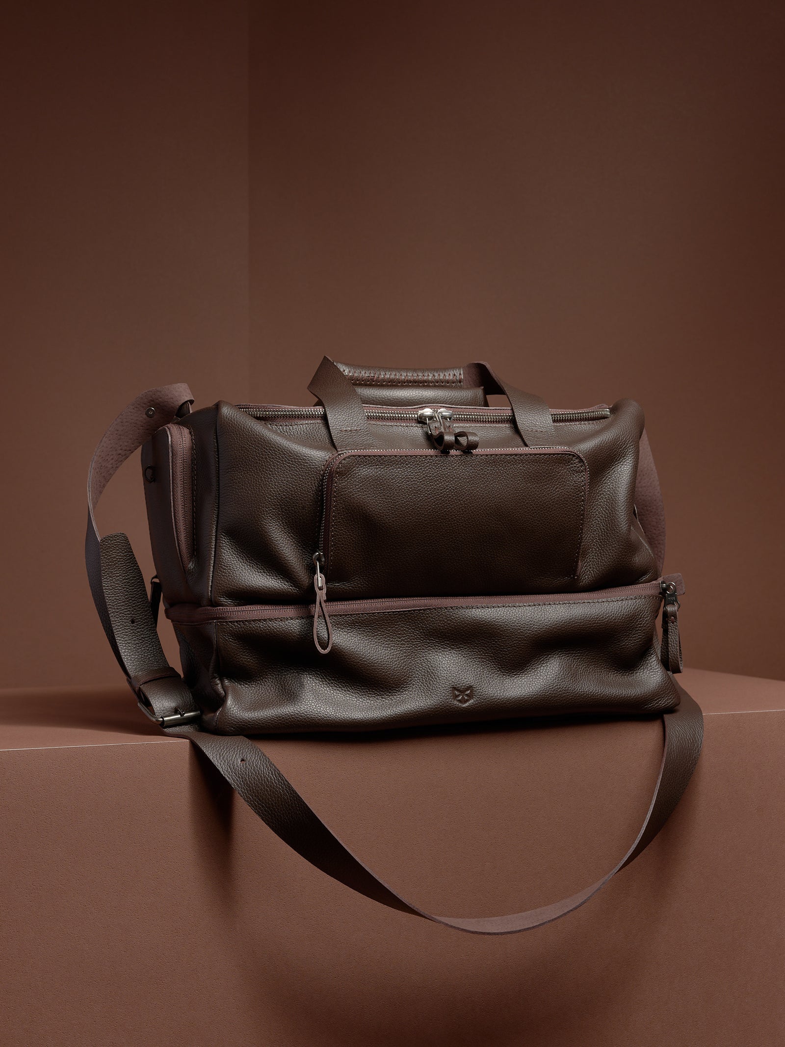 The Capra Leather Ventra Holdall, a dark brown leather duffel bag, shown on a brown background.
