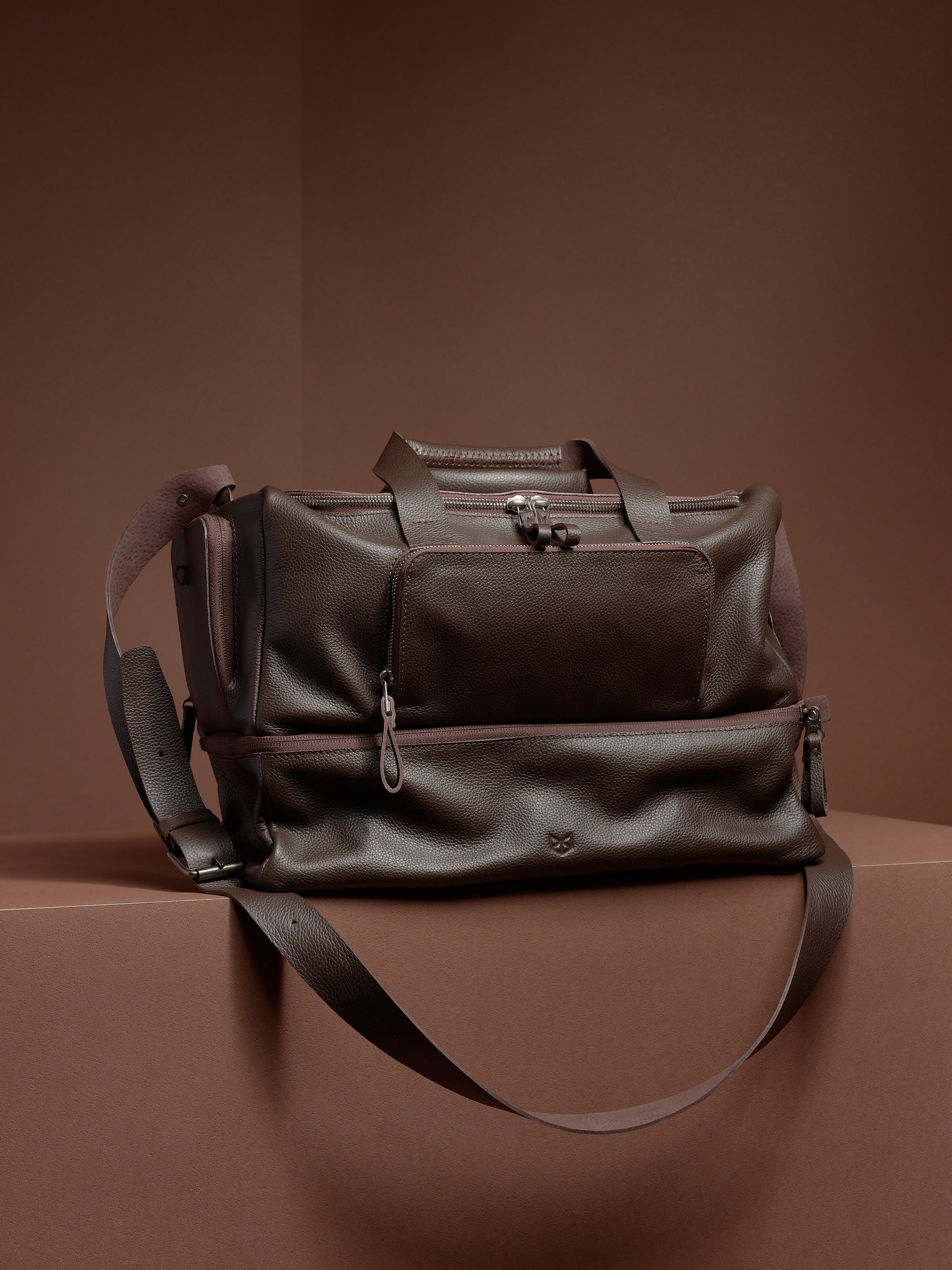 The Capra Leather Ventra Holdall, a dark brown leather duffel bag, shown on a brown background.